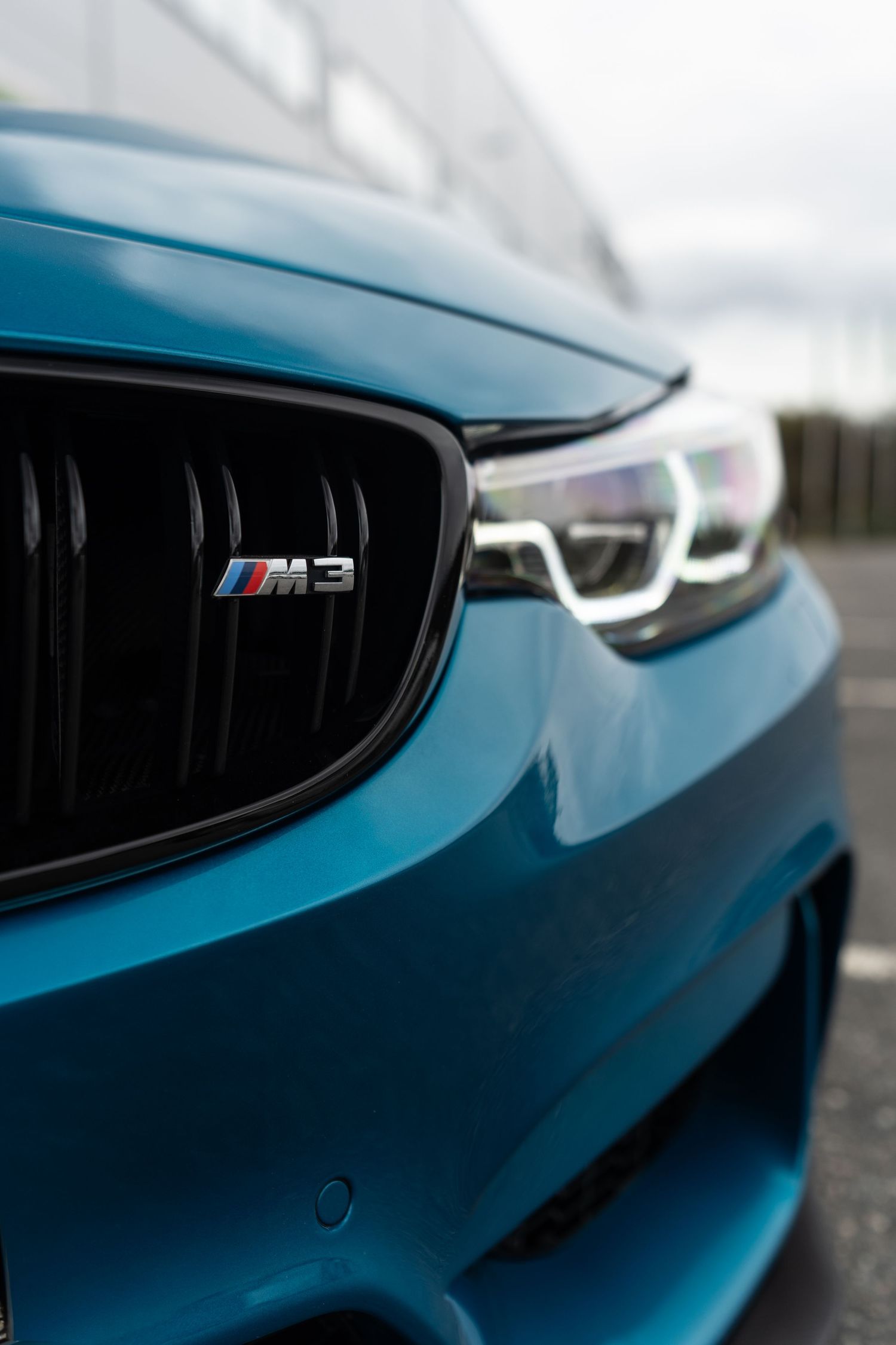 Close-up of a BMW M3 front grille and badge in metallic blue with signature LED headlights visible.
