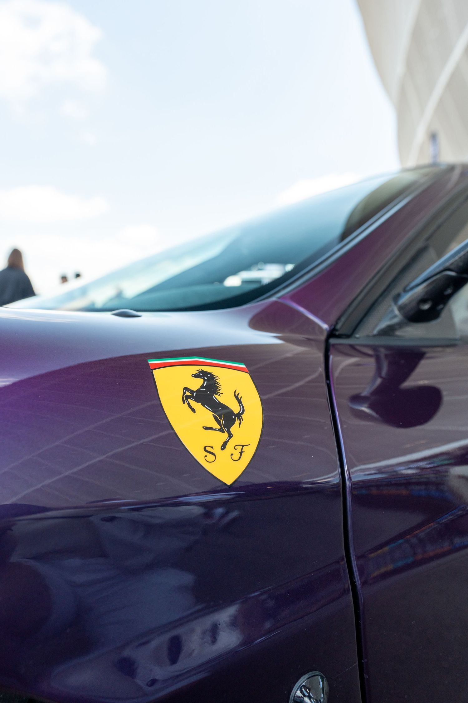 Close-up of a Ferrari badge on a dark purple car panel under bright daylight.