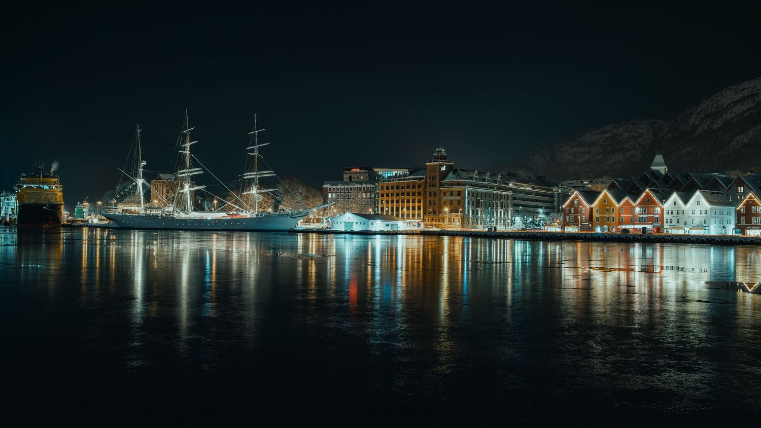 Panoramic cityscape at night showing illuminated buildings and their reflections across a calm waterfront.