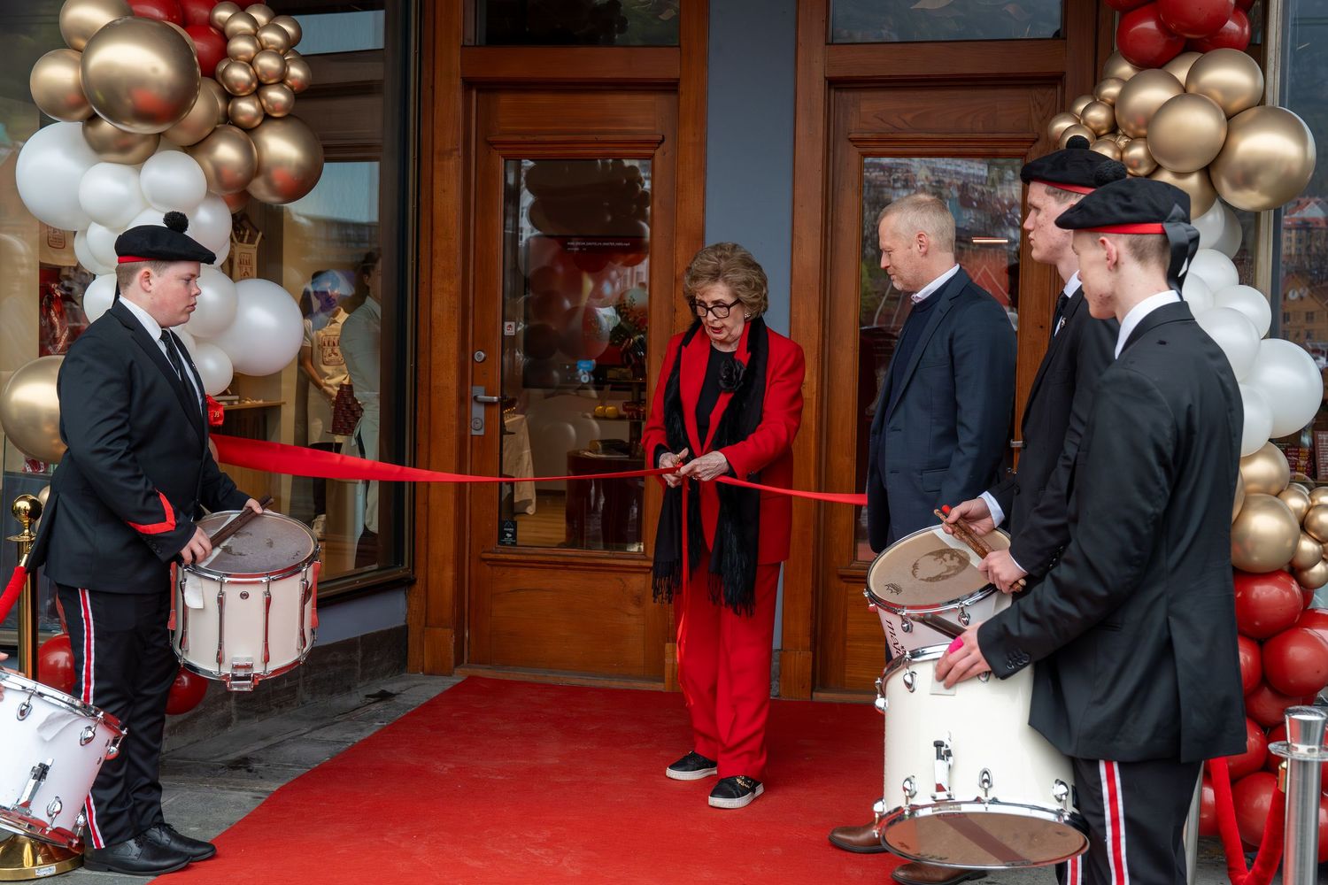 Grand opening ribbon cutting ceremony with drummers, red carpet, and balloon arch decorations outside a store entrance.