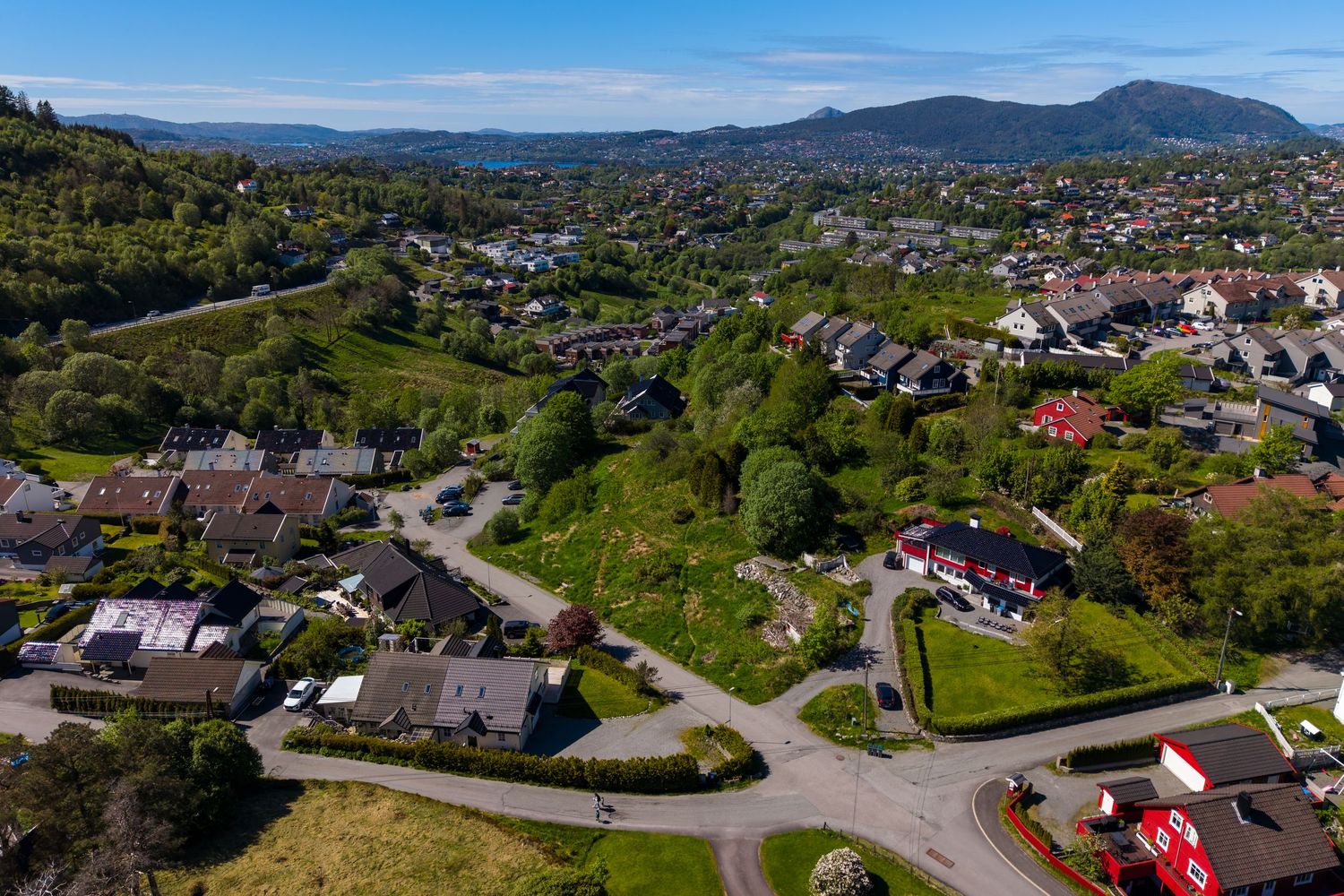 Aerial view of a green residential neighborhood with red-roofed houses, winding roads and tree-covered hills beneath distant mountains.