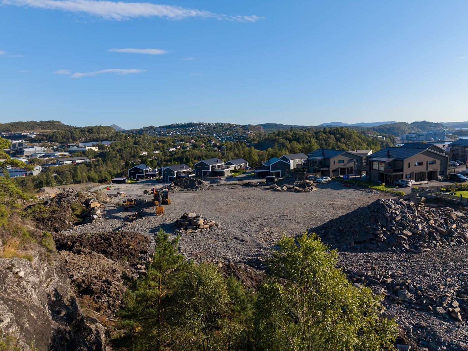 Aerial view of a cleared rocky building lot with gravel and boulders overlooking a hillside residential neighborhood under a clear blue sky.