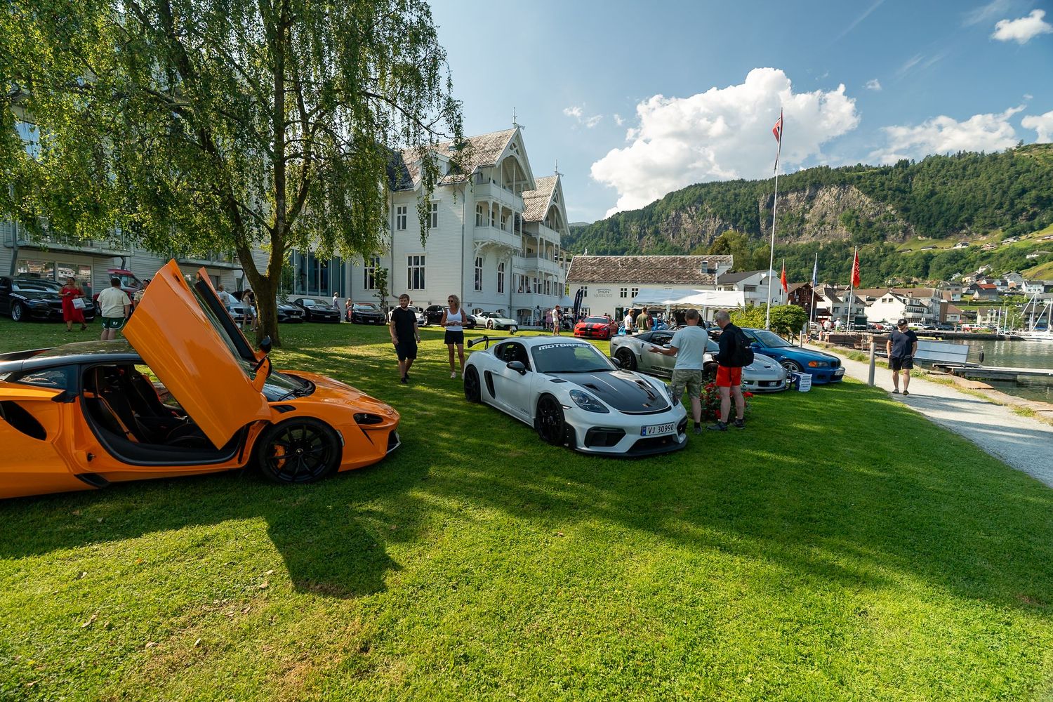 Orange McLaren with open butterfly door and white Porsche sports car parked on a grassy lawn at an outdoor car show beside a fjord village.