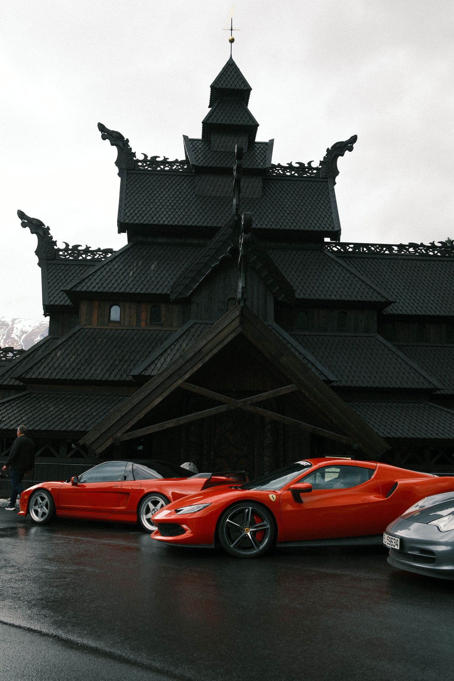 Two red Ferrari sports cars parked on wet pavement in front of a dark wooden stave church with dragon-head roof carvings.