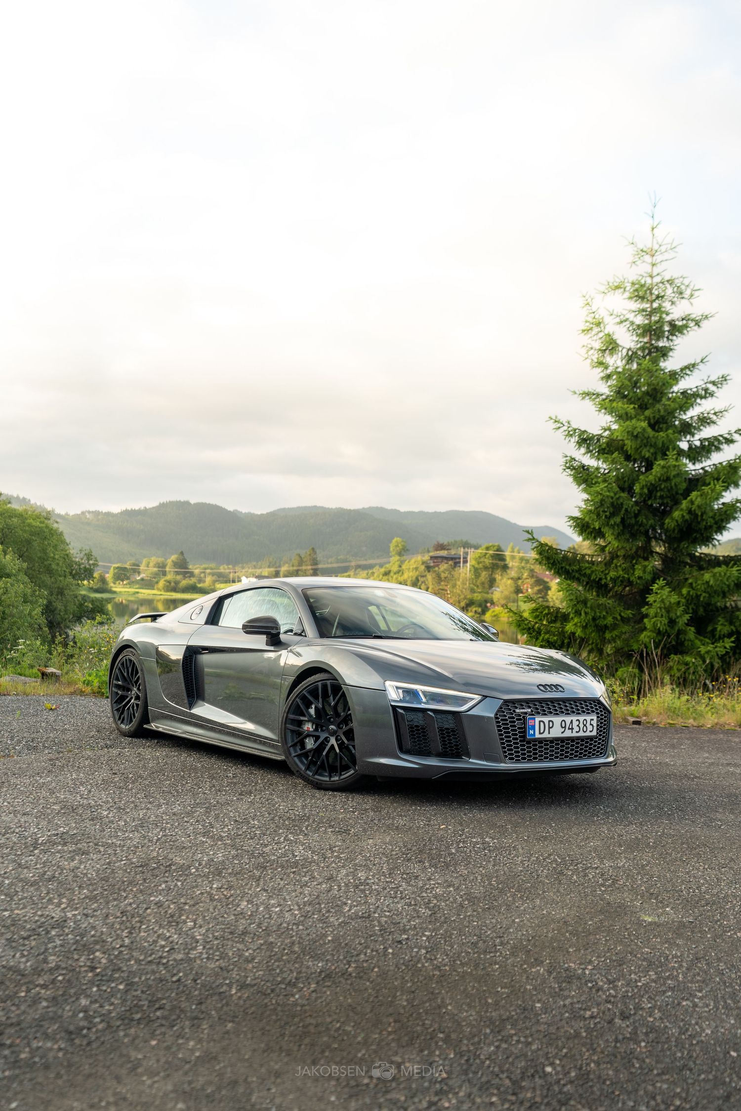 A sleek silver Audi R8 Spyder is parked on a gravel surface with mountains and trees in the scenic background.