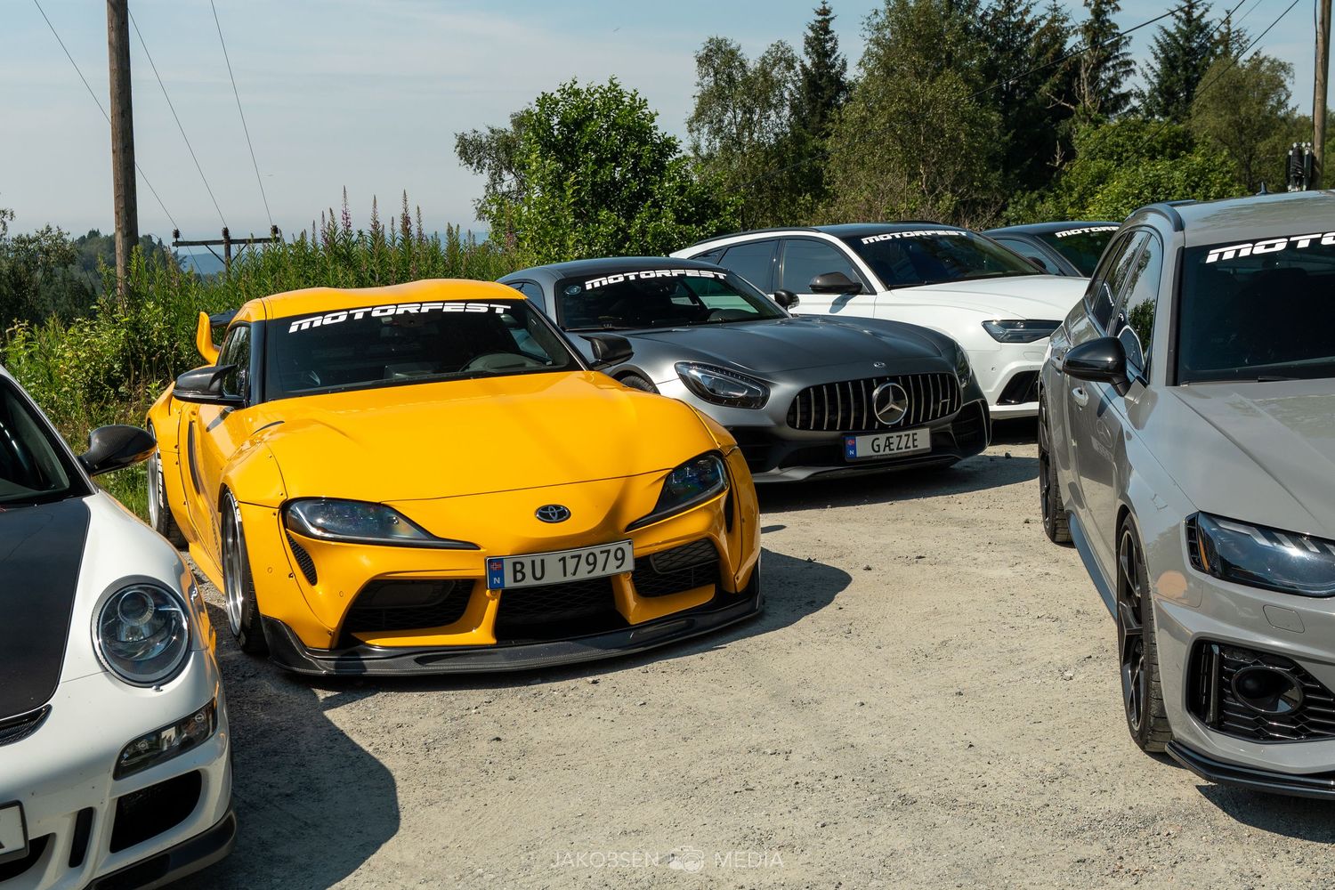 Luxury cars parked in gravel lot with mountains and trees in background.