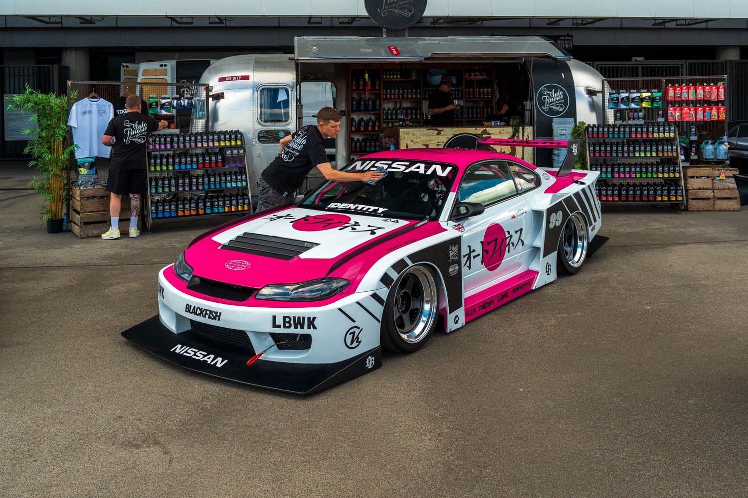 A modified white and pink race car with wide body kit and racing livery parked in front of a storefront.