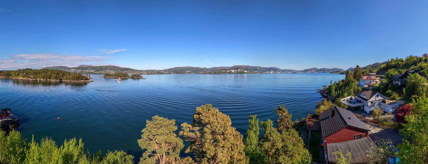 Panoramic view of a calm blue fjord surrounded by forested hills, with red and white waterfront houses and green trees under a clear sky.