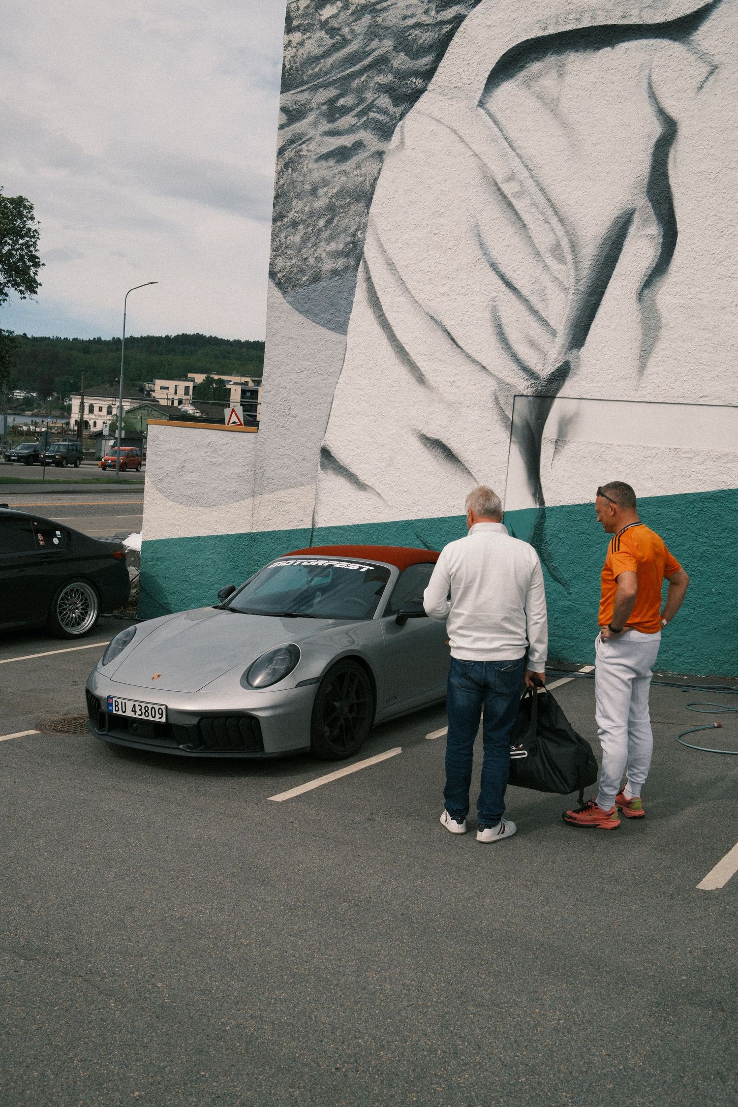 Two men stand beside a silver Porsche convertible in a parking lot in front of a large black and white mural on a teal wall.