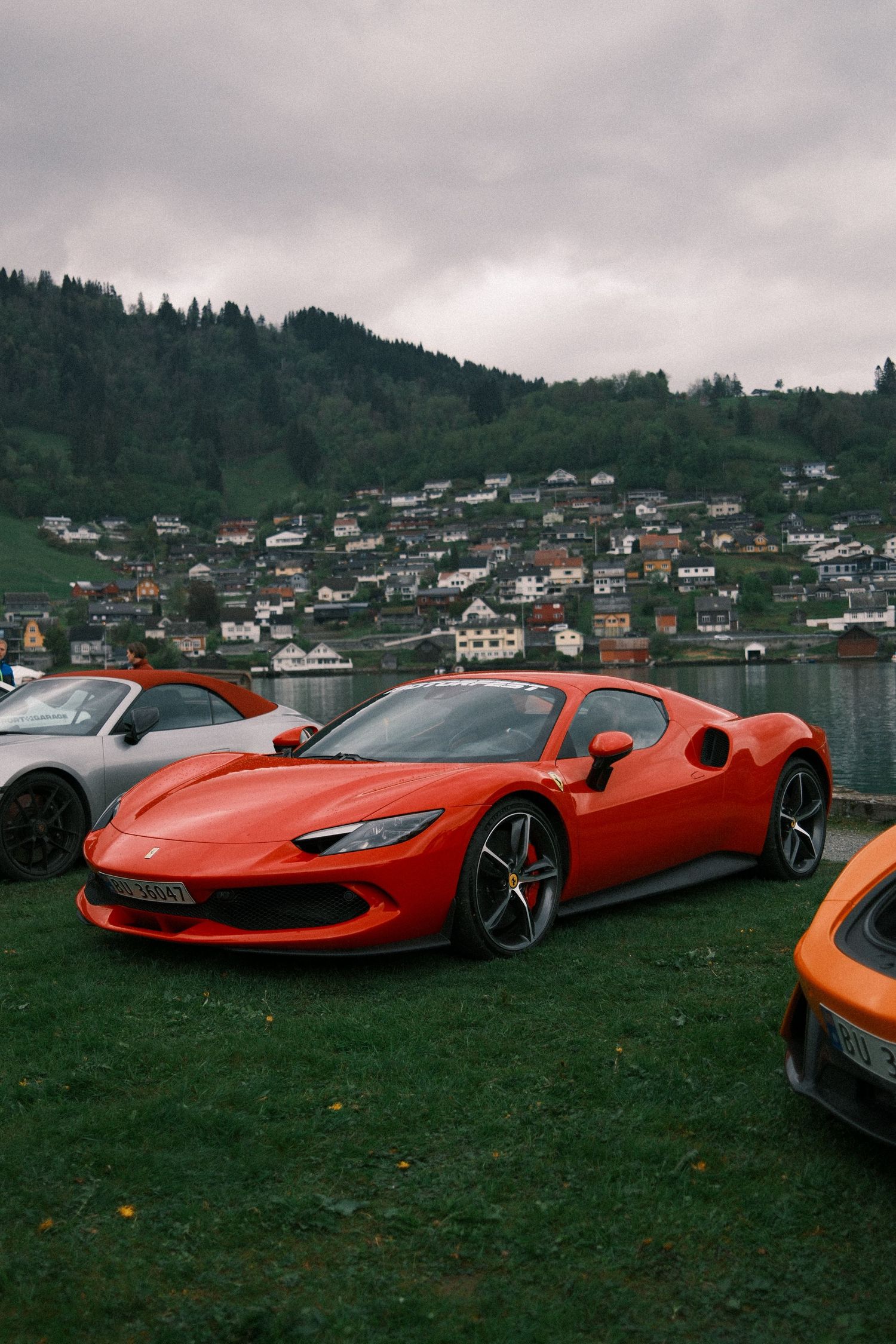 Red Ferrari 296 GTB parked on a grassy lakeside lawn with a colorful hillside town and forested mountains under an overcast sky.