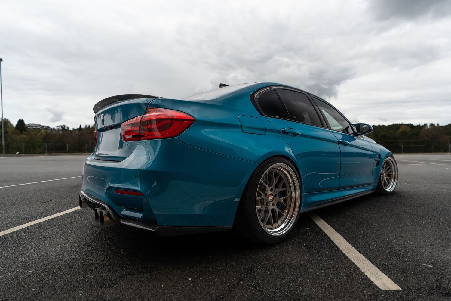 Custom modified BMW M3 F80 in metallic teal blue with aftermarket wheels parked on an empty highway.