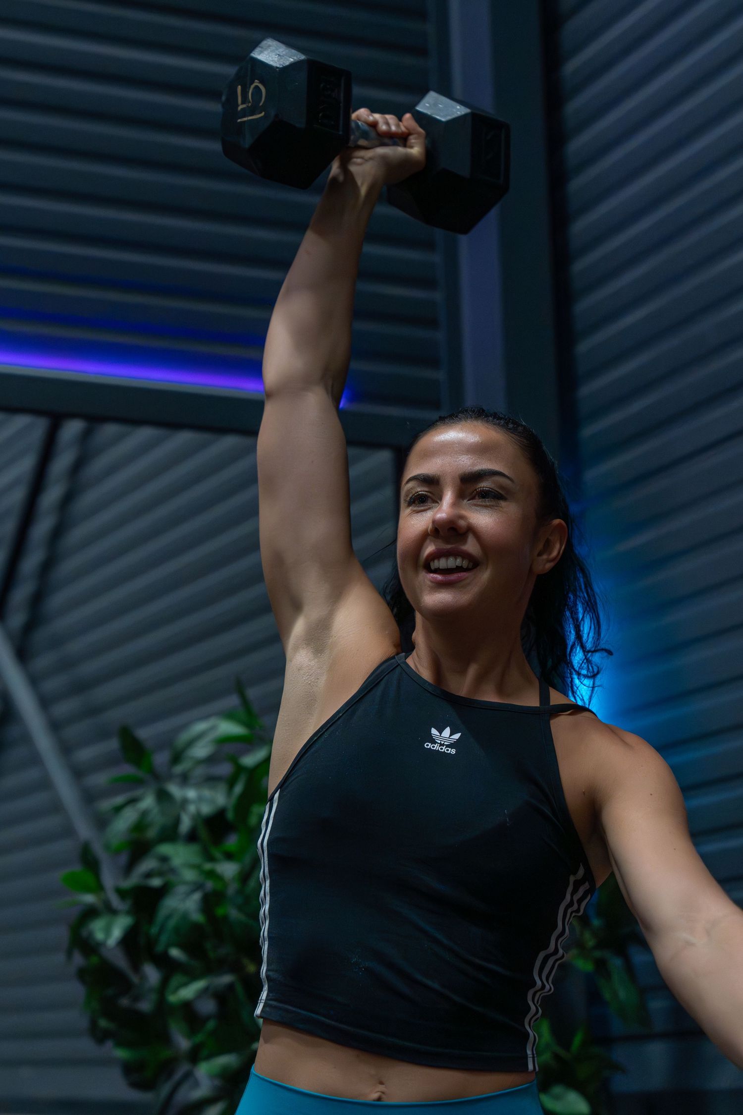 Athletic person in black Adidas crop top lifting a dumbbell overhead during workout session with blue lighting.