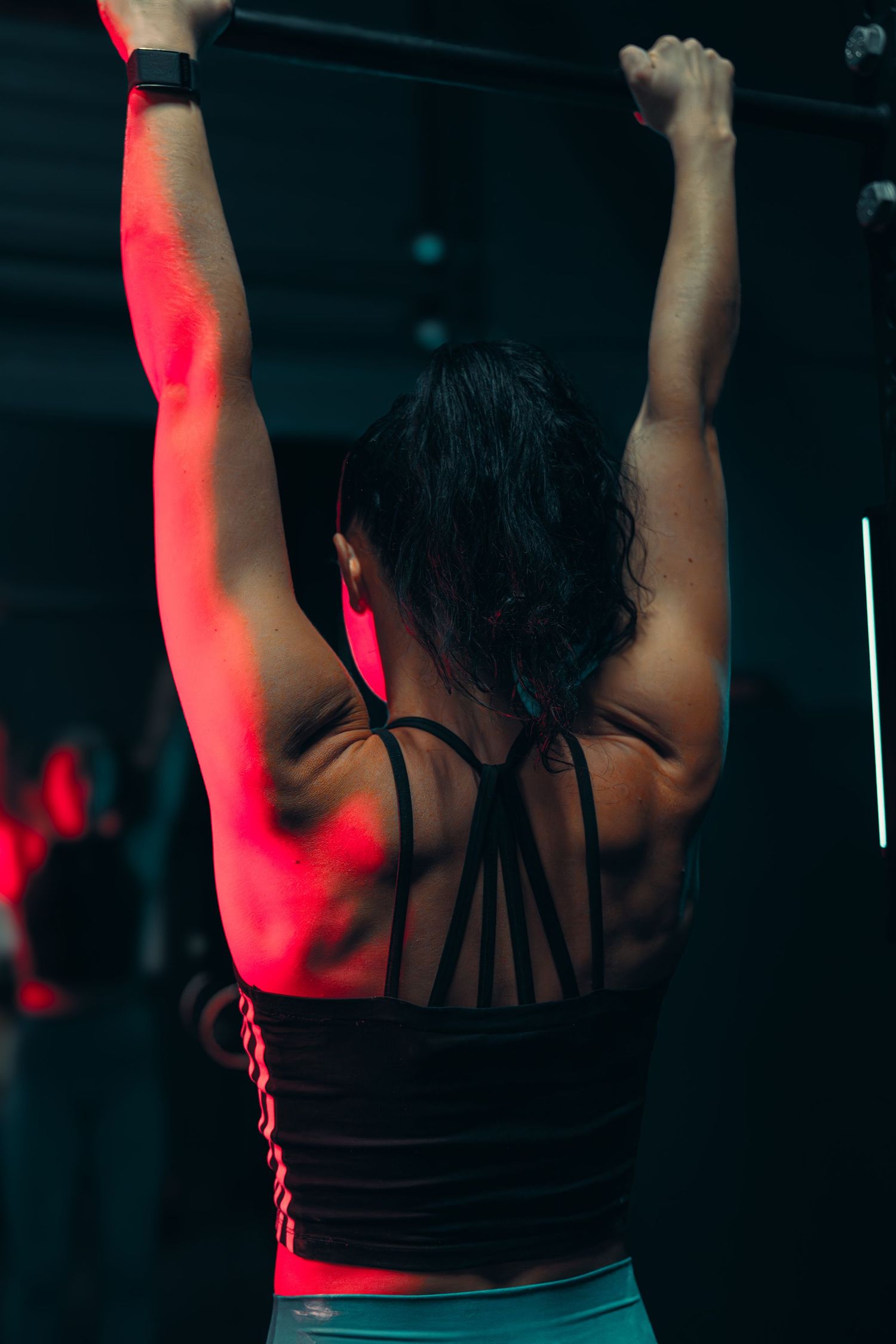 Athletic person in strappy tank top doing pull-ups under red lighting.