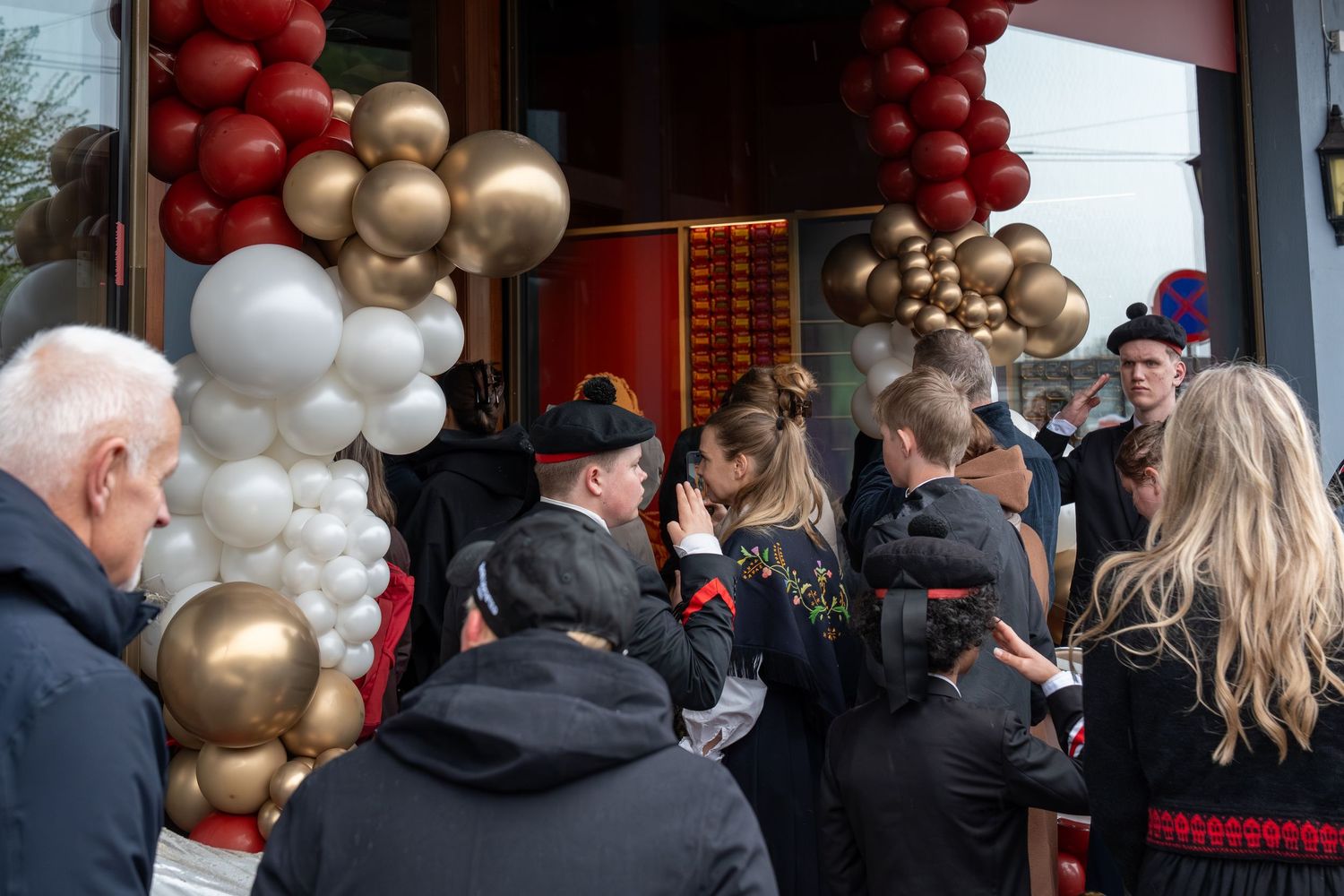 Crowd gathers outside a storefront decorated with red, white and gold balloon garlands for a festive grand opening celebration.