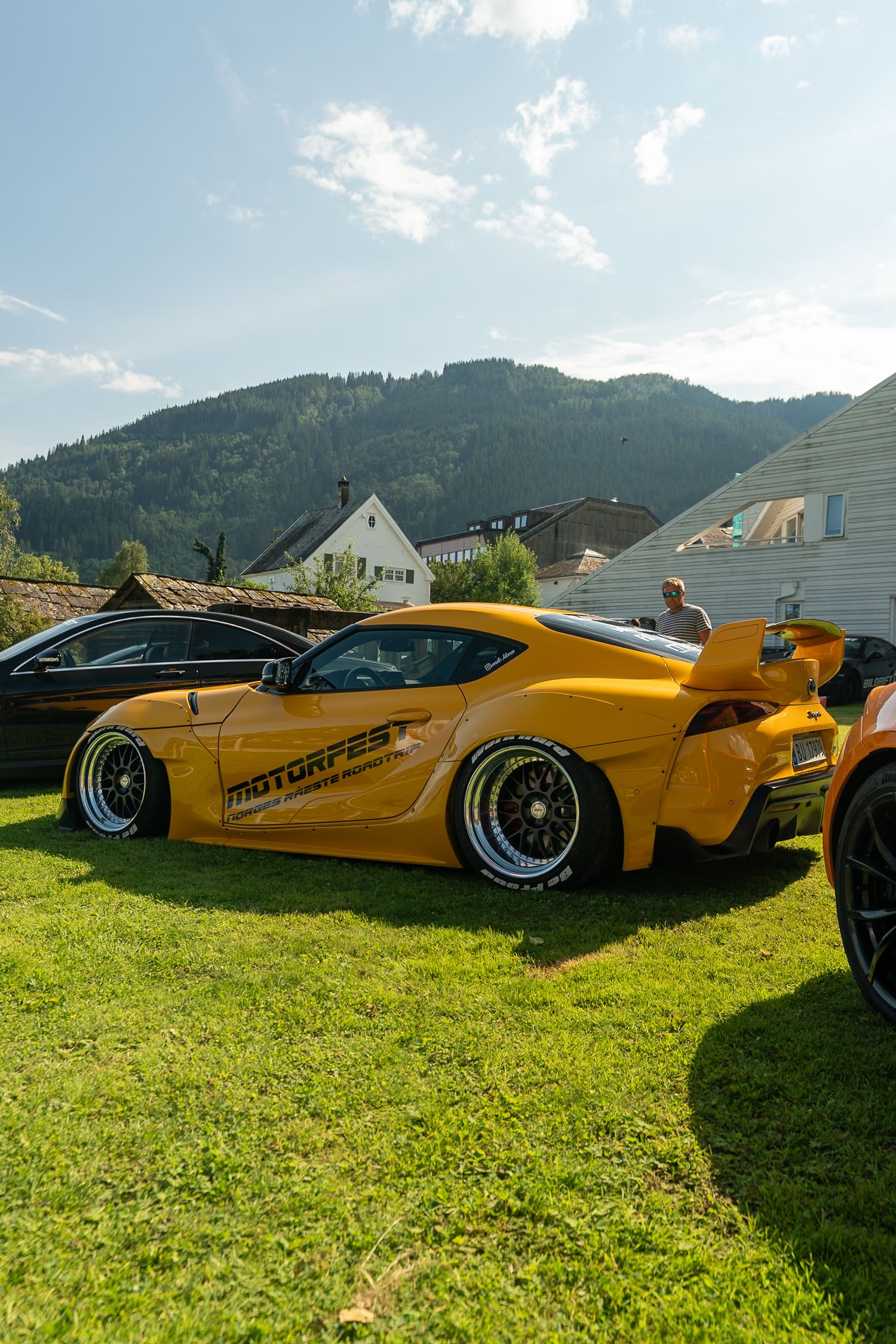 Orange widebody Toyota Supra with large rear wing and black mesh wheels parked on green grass at a car show with a mountain backdrop.