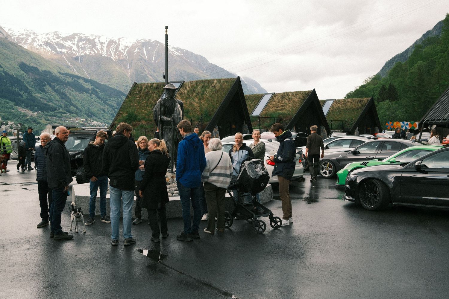 Group of tourists gathers on a wet plaza near parked cars and sod-roofed huts with snow-capped mountains rising in the background.