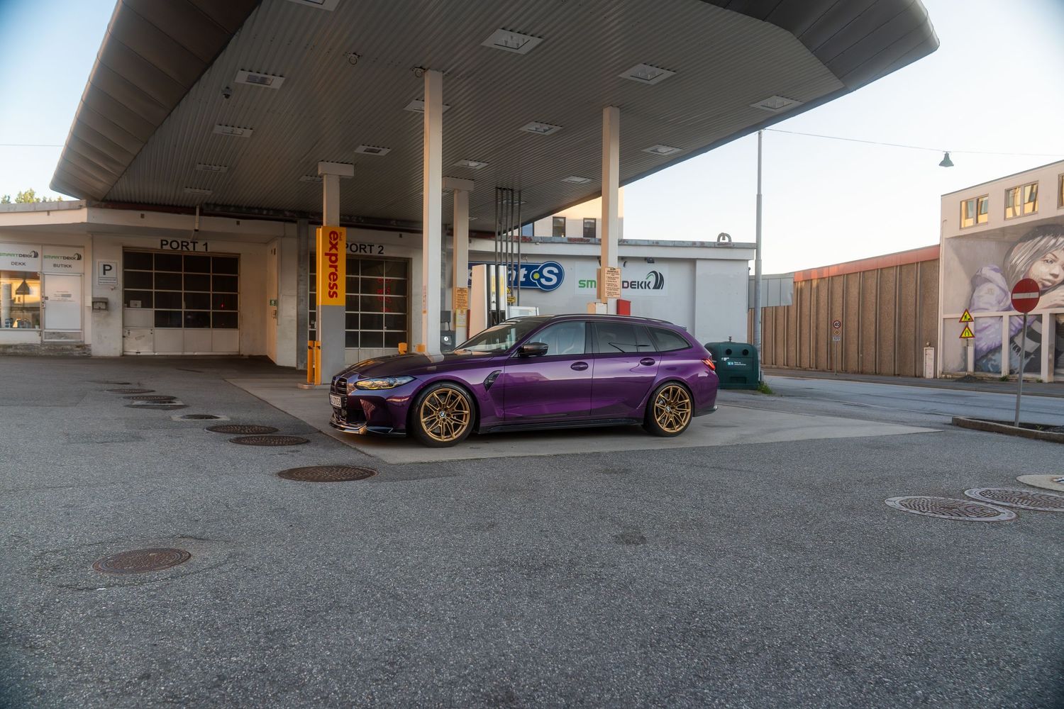 Wide angle view of a modified purple BMW M3 parked under a covered concrete overhang at dusk.