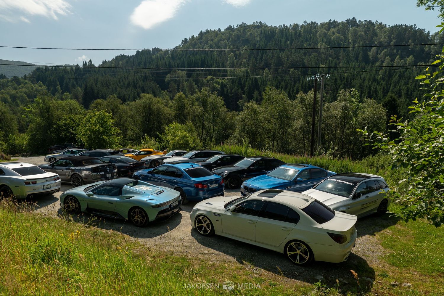 Collection of luxury cars parked on a hilltop overlook with forested mountains in the background.