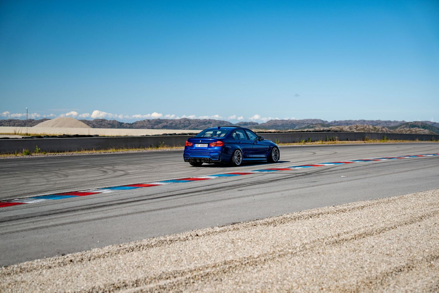 Blue sports car speeding down a racetrack with mountains visible in the background against a bright blue sky.