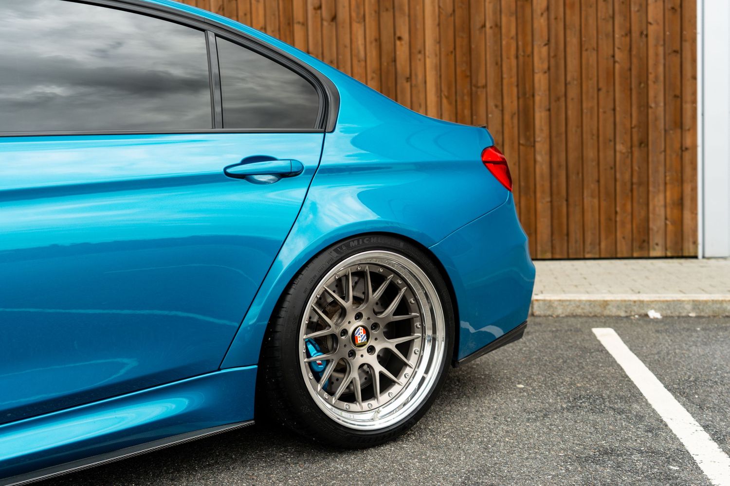 A bright blue BMW sports sedan with silver multi-spoke wheels sits against a wooden wall in a parking space.