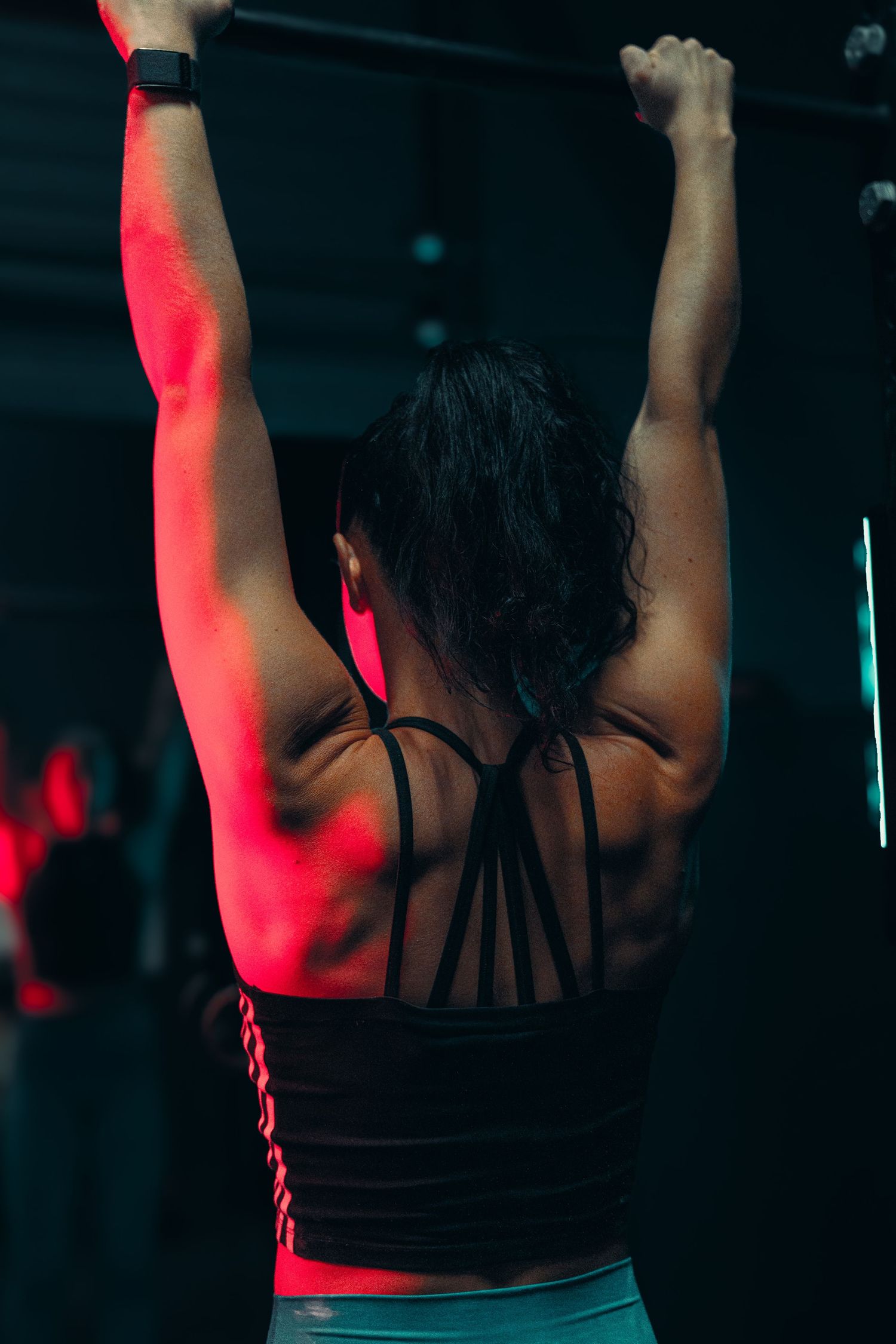 Back view of an athlete in black strappy top raising arms overhead with red lighting in dark gym setting.