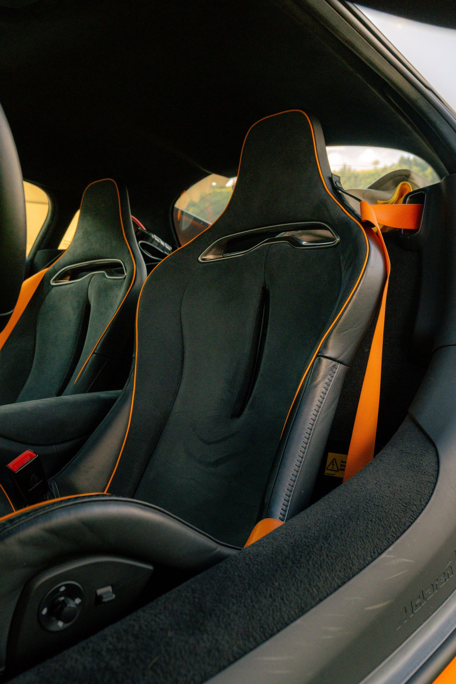 McLaren sports car interior featuring black suede bucket seats with orange contrast stitching and matching orange racing harness seatbelts.