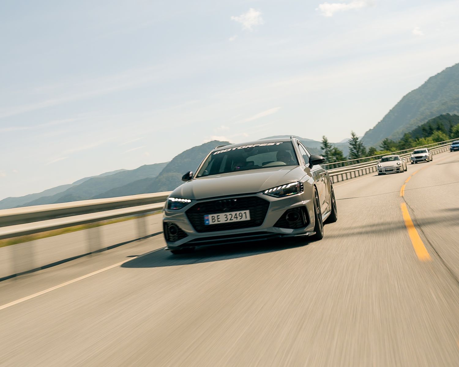 Gray Audi RS4 Avant wagon driving along a sunlit mountain road with scenic peaks and cars following behind.