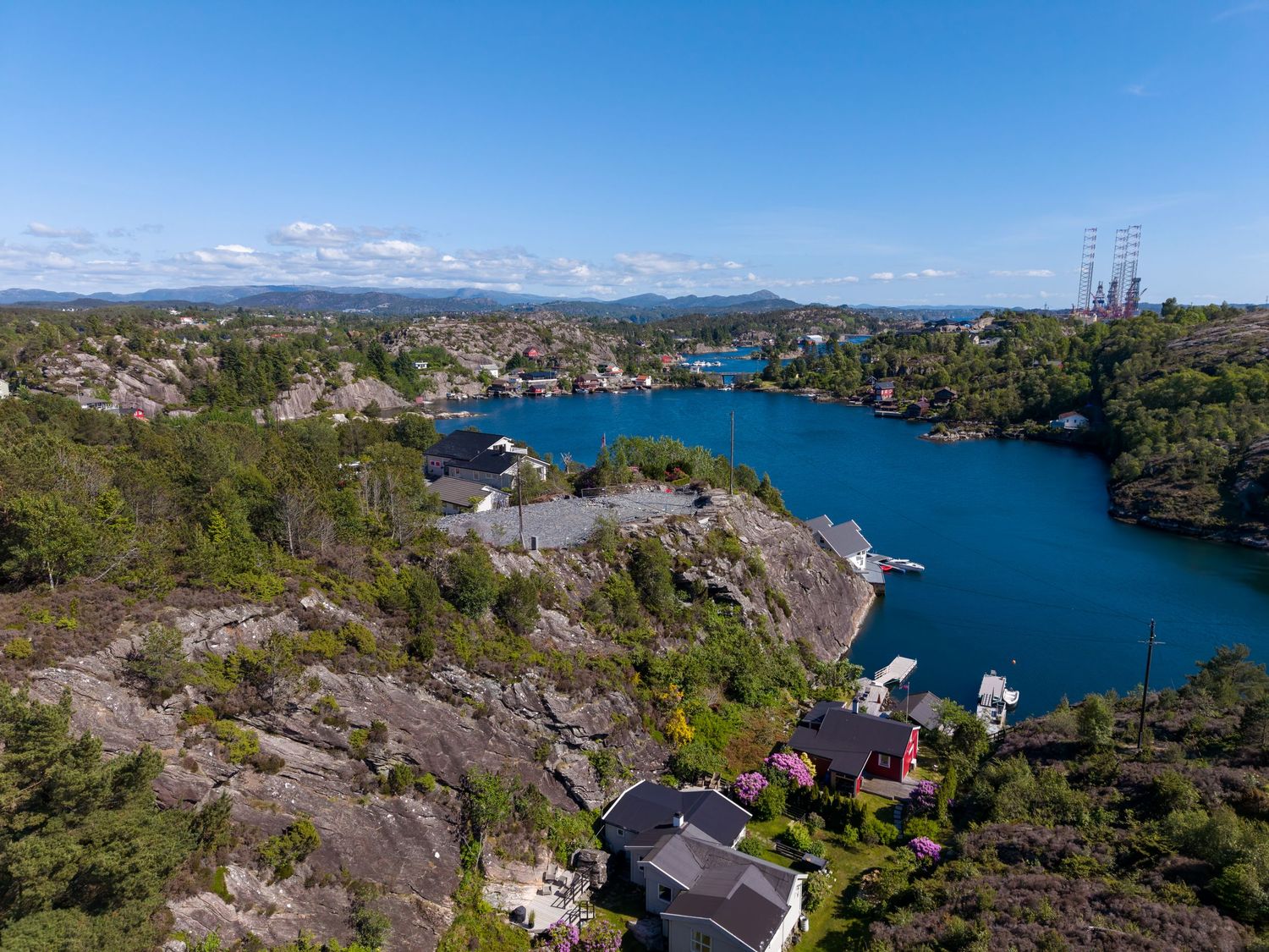 Aerial view of a deep blue fjord winding past rocky, tree-covered hillsides with scattered coastal homes under a clear sky.