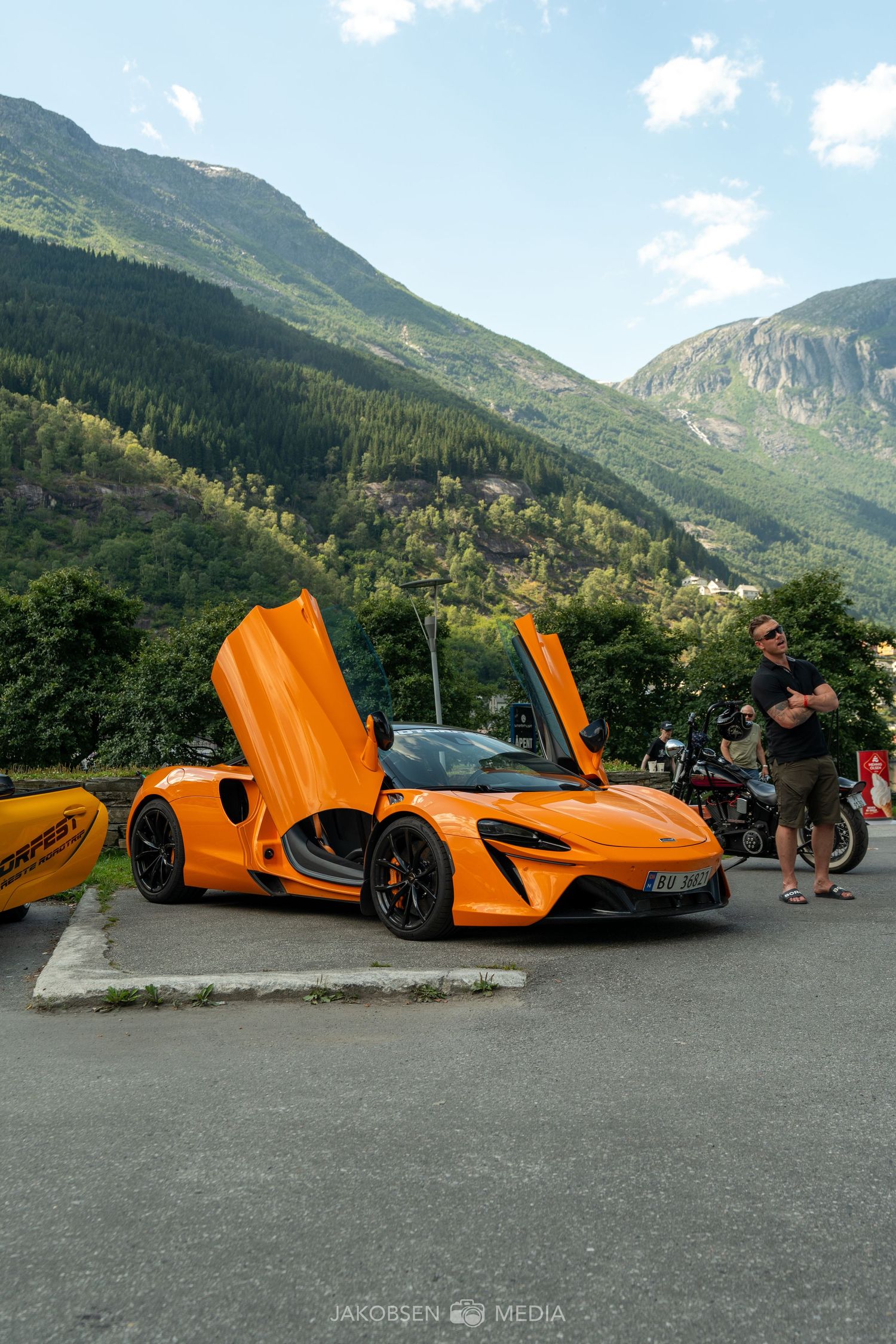 Orange McLaren sports car with butterfly doors open parked in scenic mountain landscape.