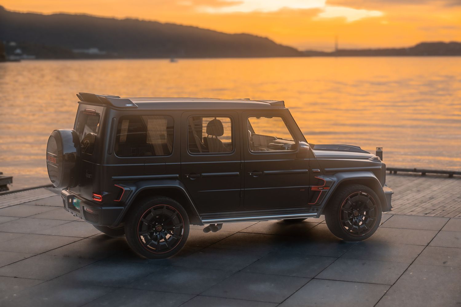 Black Mercedes-AMG G-Wagon SUV parked against dramatic orange sunset at waterfront location.