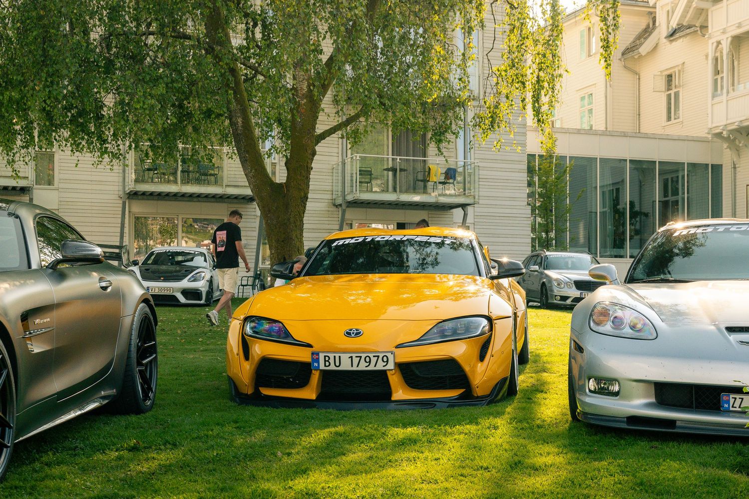 Yellow Toyota Supra with Norwegian plate parked on green grass between sports cars beside a historic building and a leafy tree.