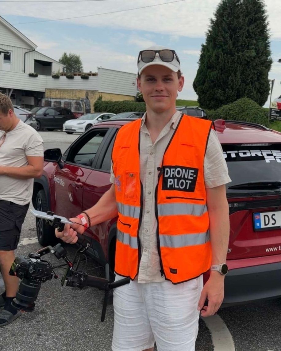 Man in orange high-visibility vest labeled Drone Pilot holds a drone controller beside a red car in a sunny parking lot.