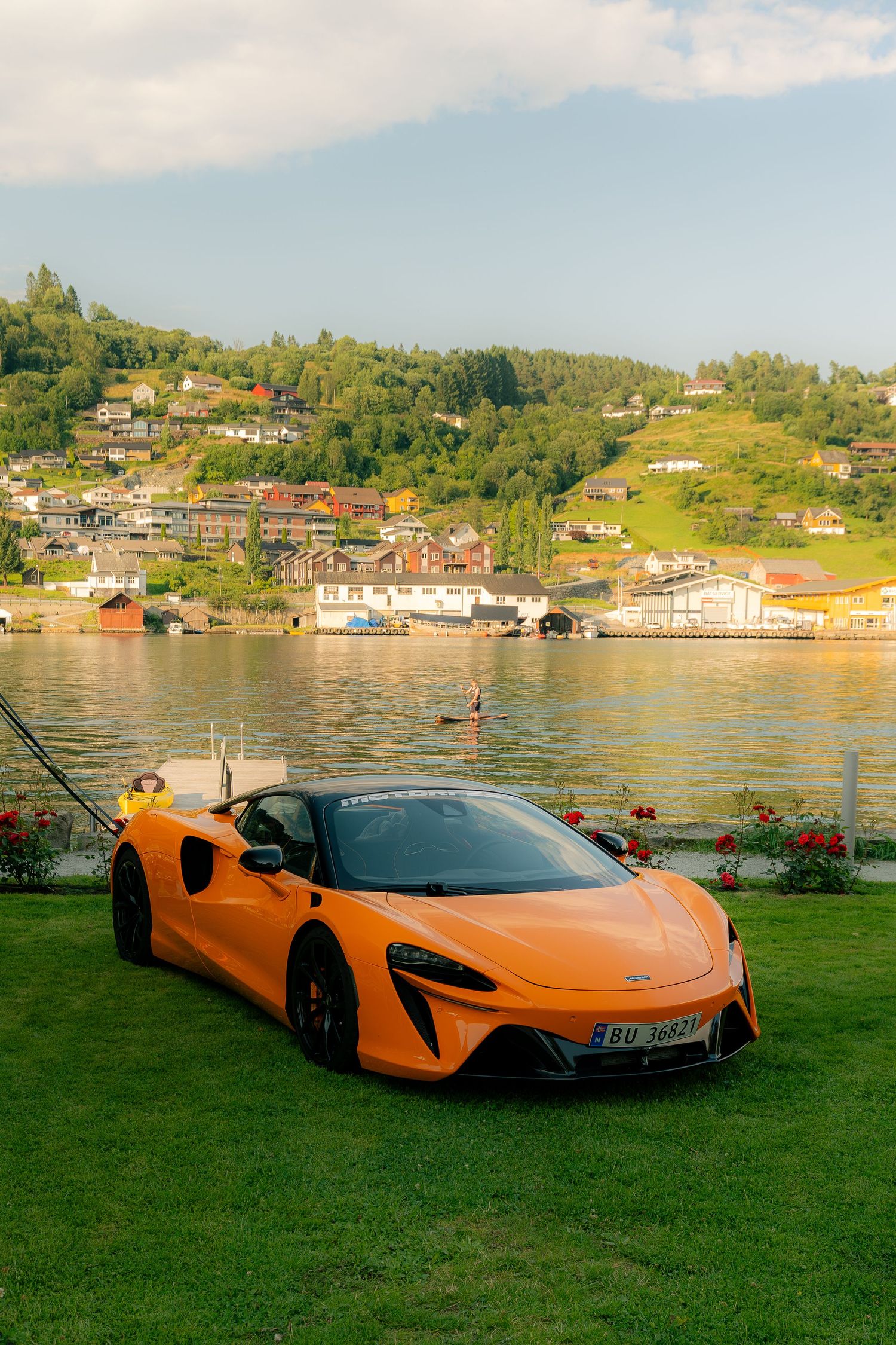 Orange McLaren Artura supercar parked on green grass beside a scenic Norwegian fjord with hillside village in background.