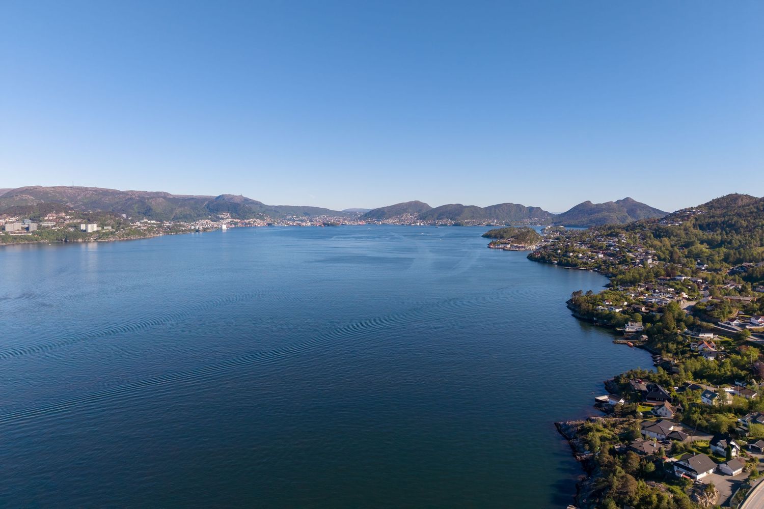 Aerial view of a calm blue fjord bordered by green hills, waterfront homes, and distant mountains under a clear sky.