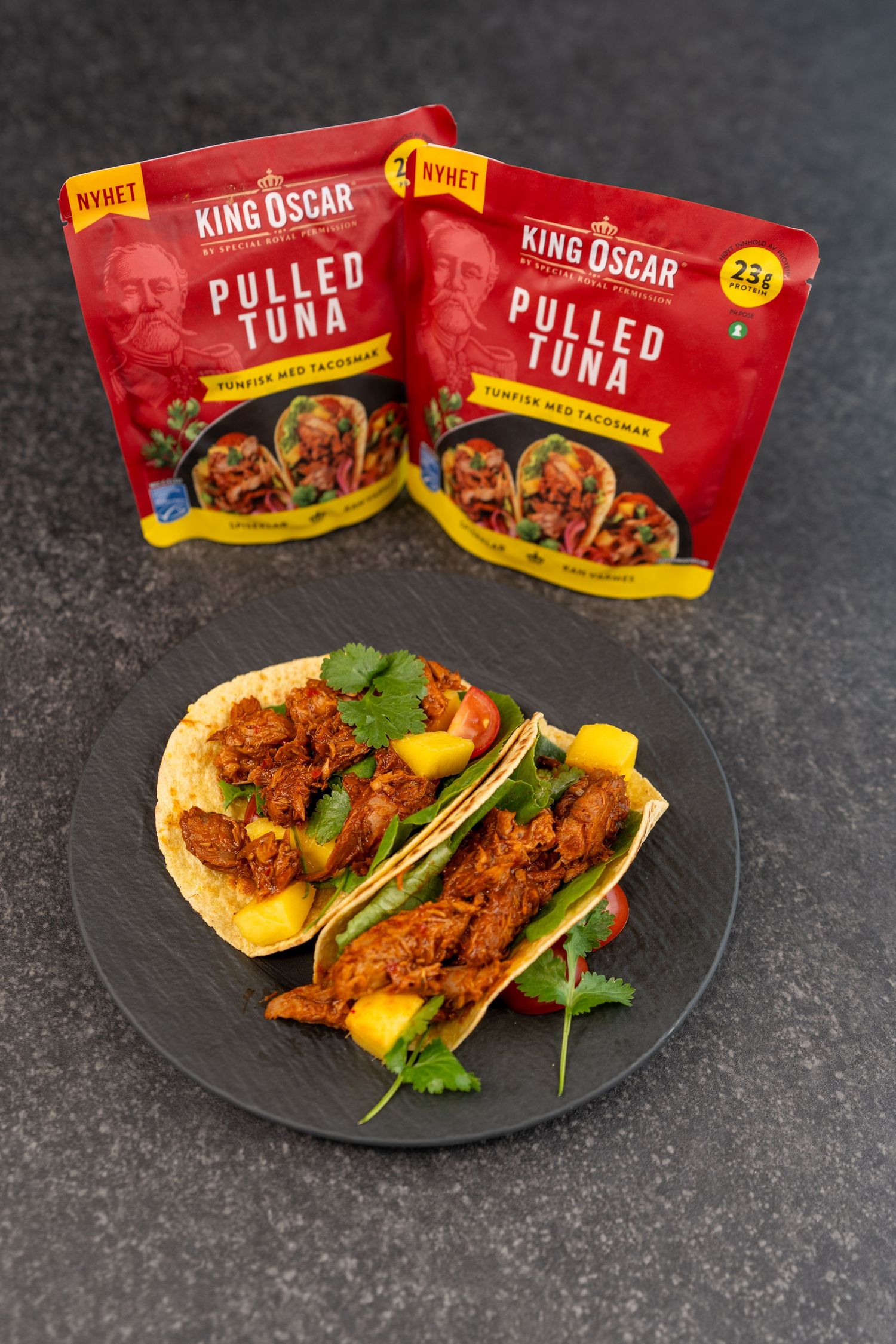 Two King Oscar Pulled Tuna pouches behind a black plate of tacos filled with tuna, mango, fresh cilantro and tomato.