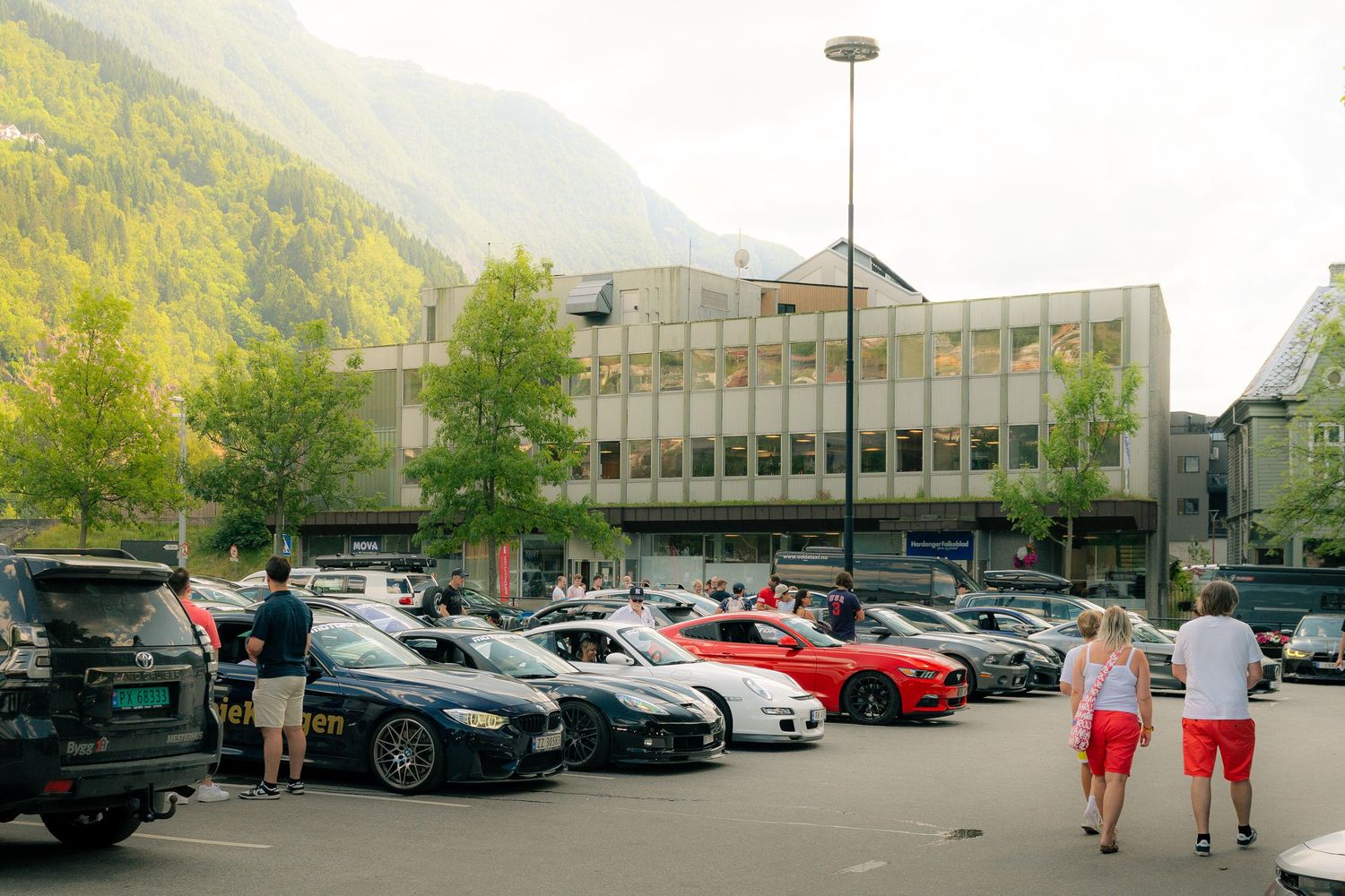 Group of sports cars parked in a lot near a modern building with people walking, set against green alpine mountains and trees.