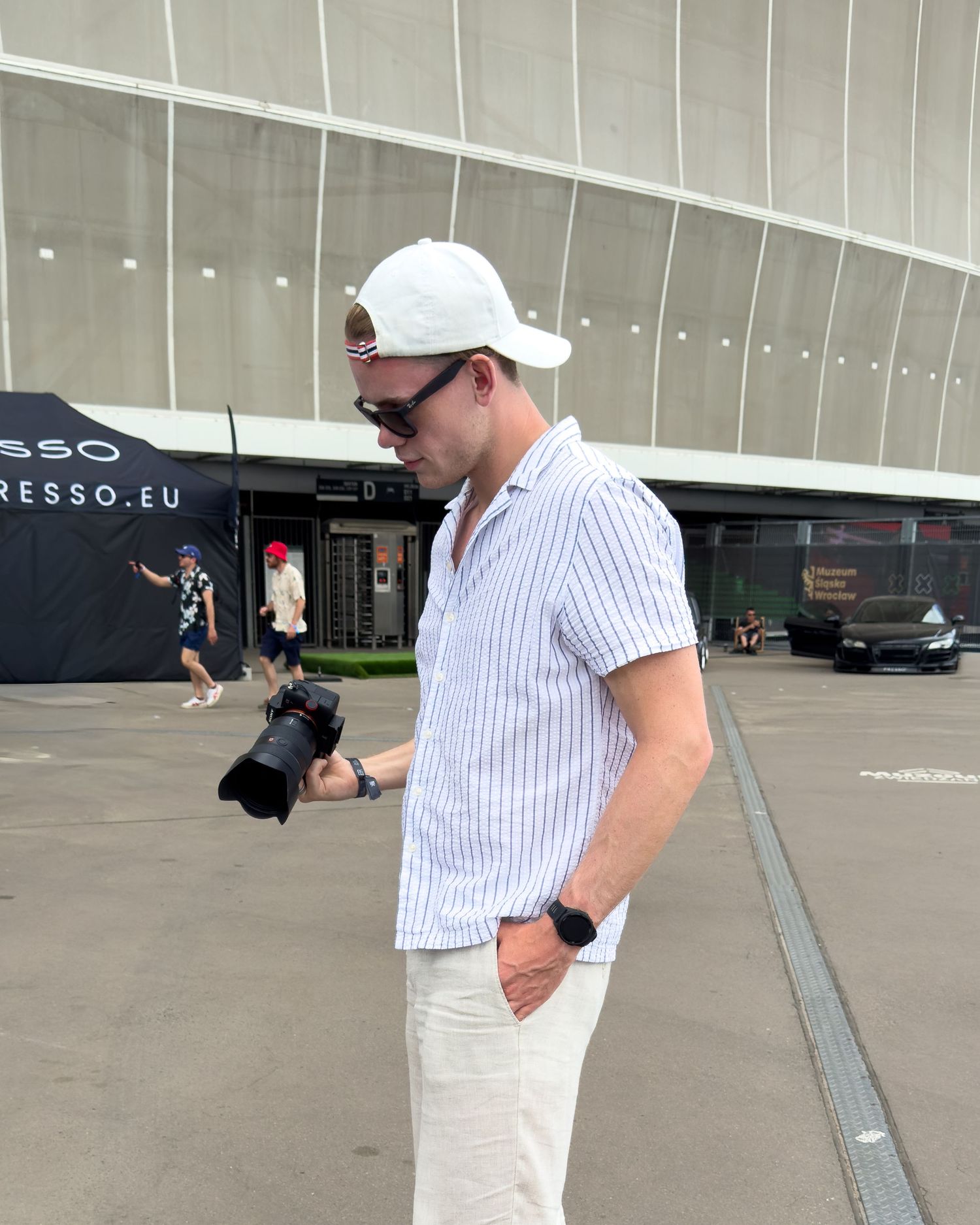 Man in white cap and striped shirt holding a camera outdoors near a modern building.
