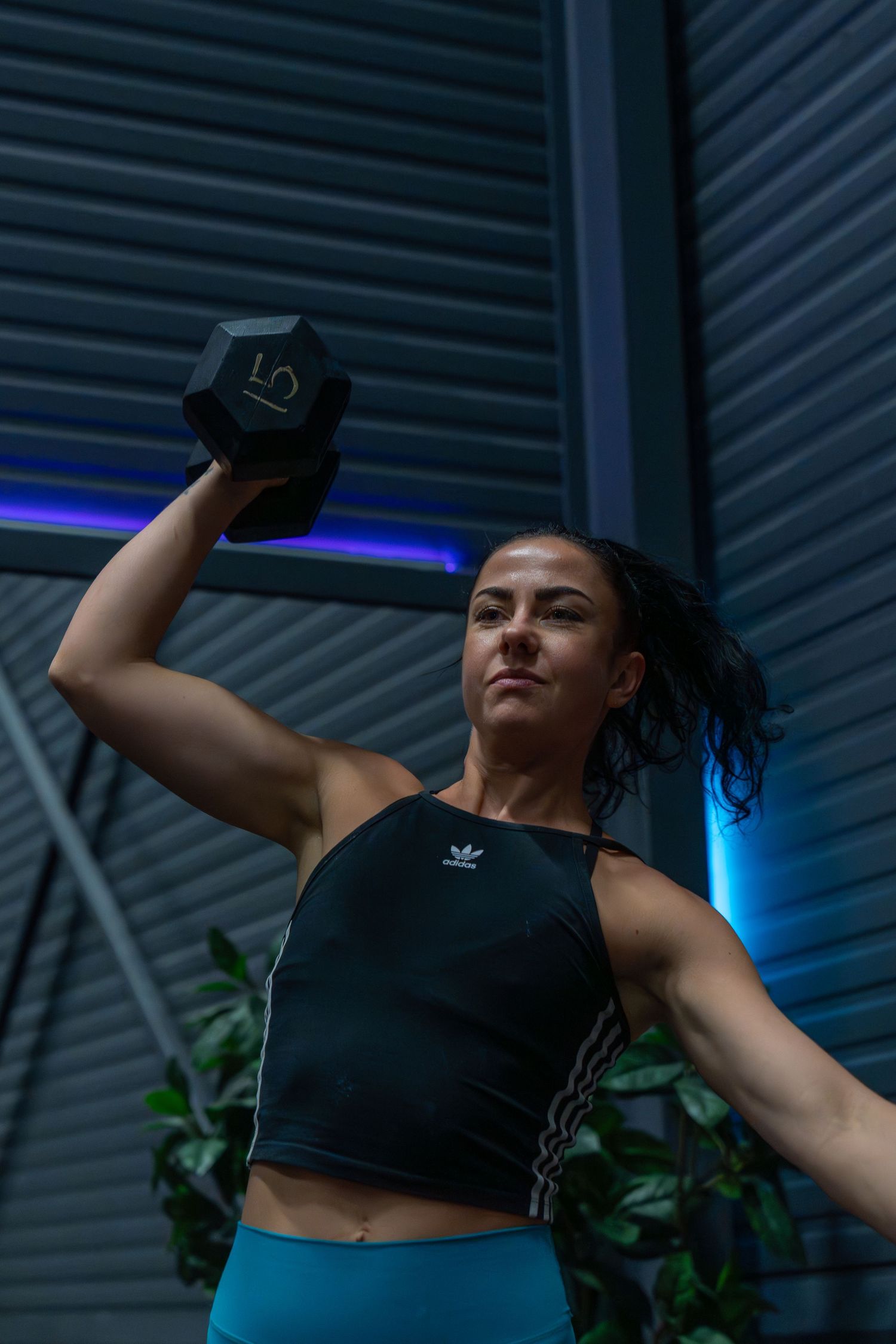 Athletic woman in Adidas crop top performs dumbbell exercise with intense focus against industrial backdrop with blue lighting.