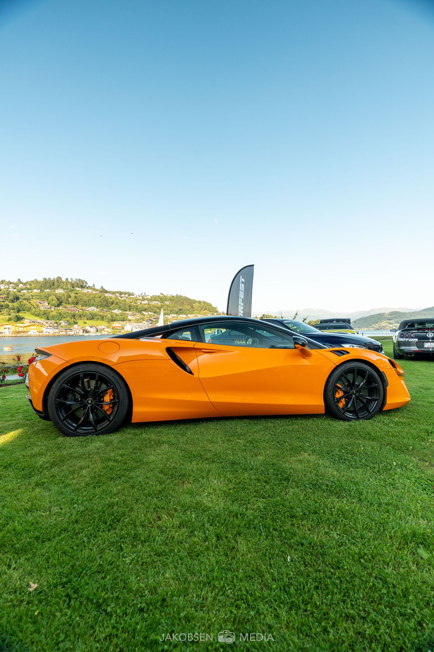 McLaren sports car in bright orange sits on green grass during an outdoor showcase event.