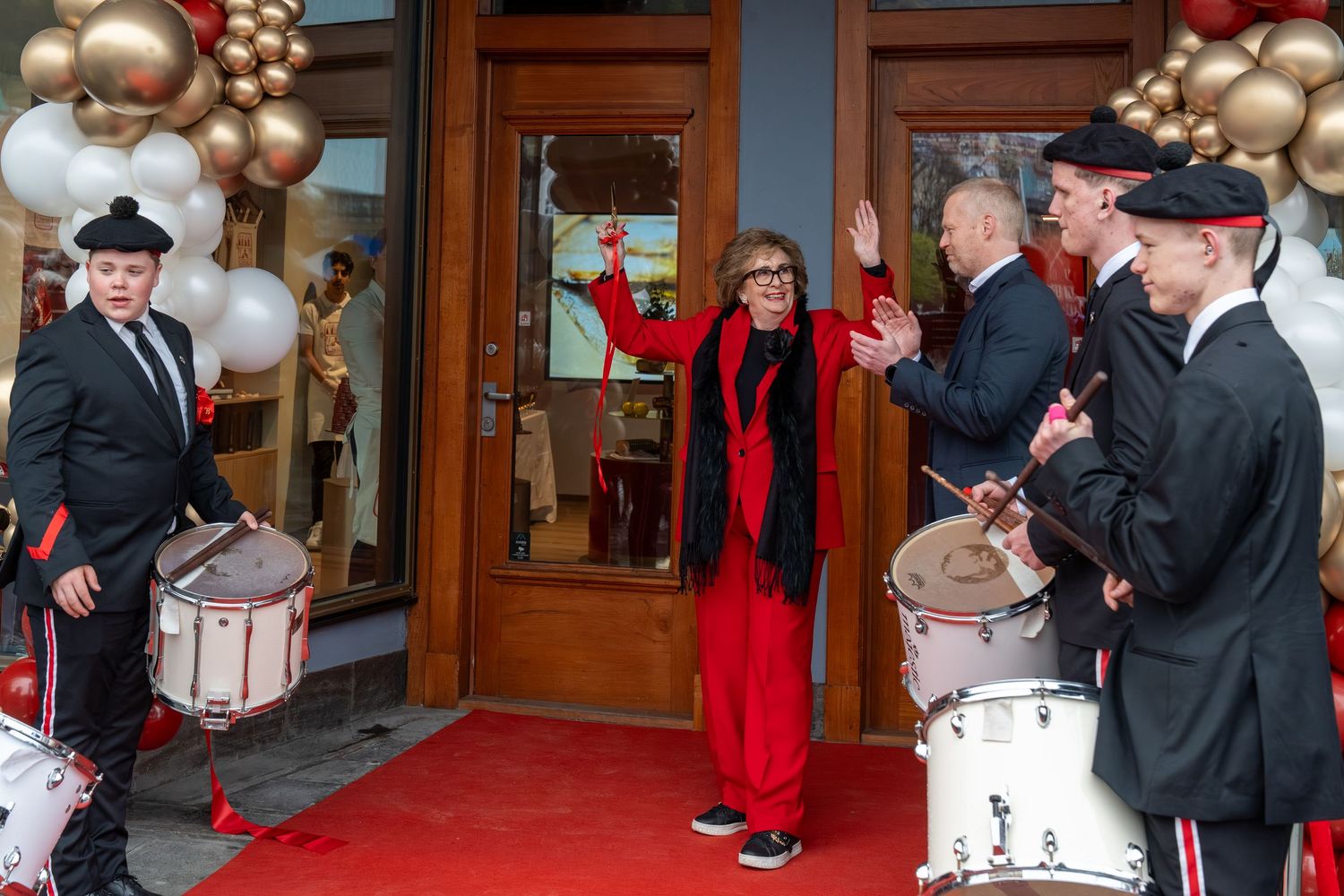 Woman in red outfit cheers on a red carpet at a storefront grand opening with drummers in uniform and balloon garlands.