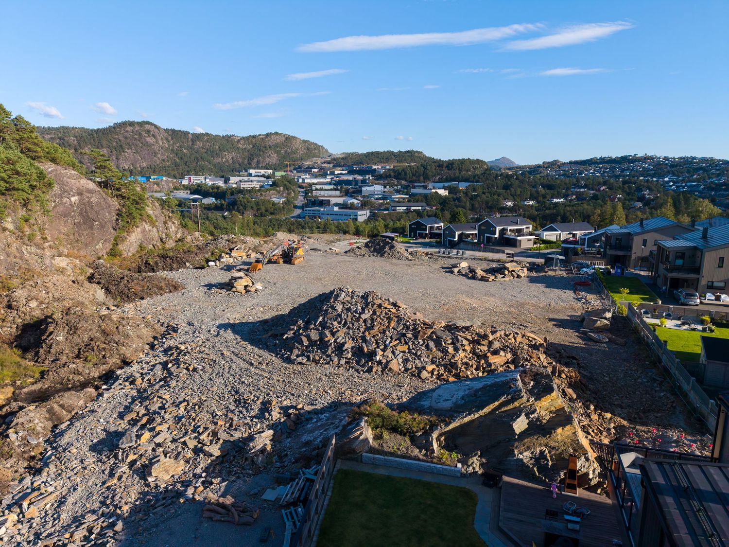 Aerial view of a rocky construction site with excavated rubble beside a hillside residential neighborhood on a clear sunny day.