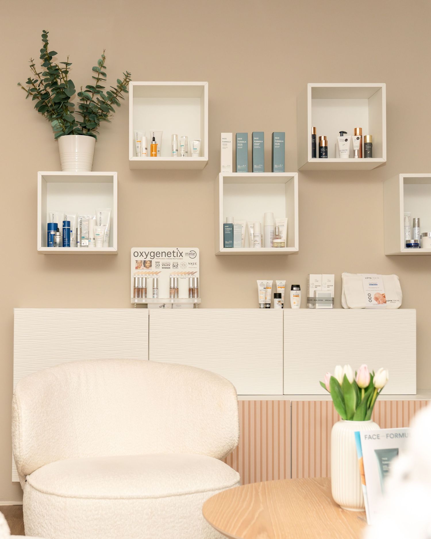 Cozy skincare boutique corner with white cube shelves of beauty products, an Oxygenetix display, a cream armchair, and fresh tulips.