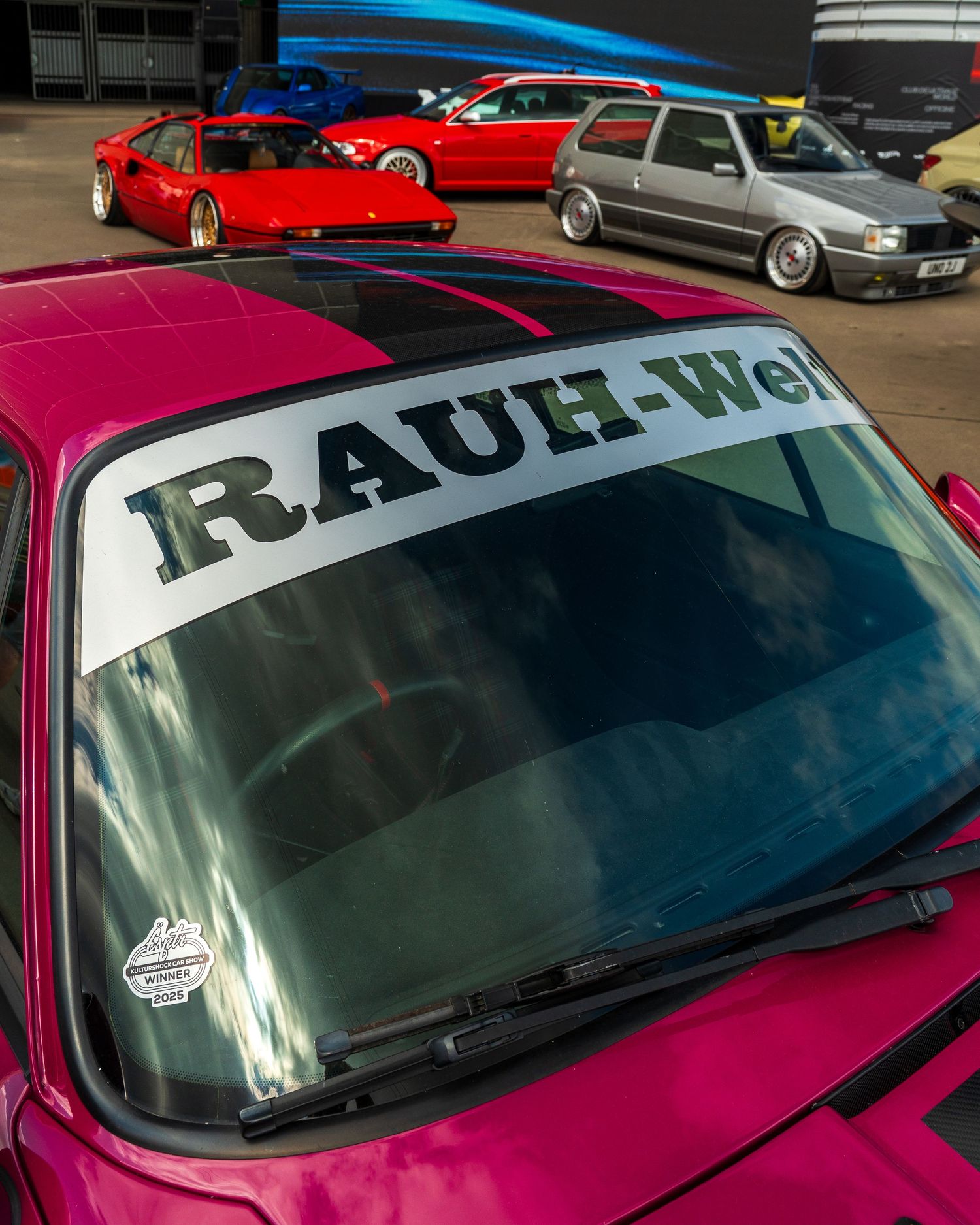 Pink car with RAUH-Welt windshield banner displayed among other modified vehicles.