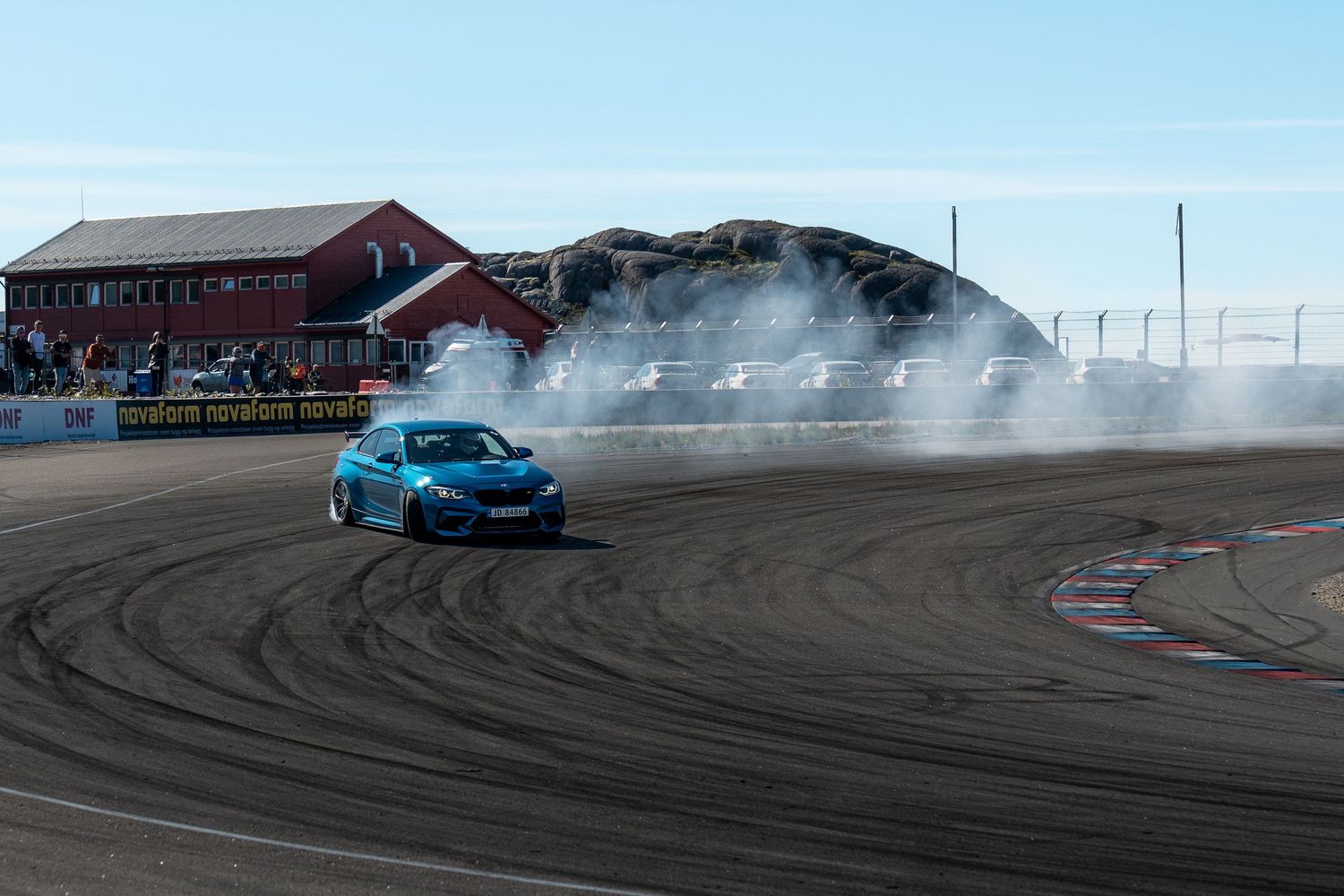 A bright blue sports car performs a drift, leaving tire marks and smoke on a racetrack with red buildings in the background.