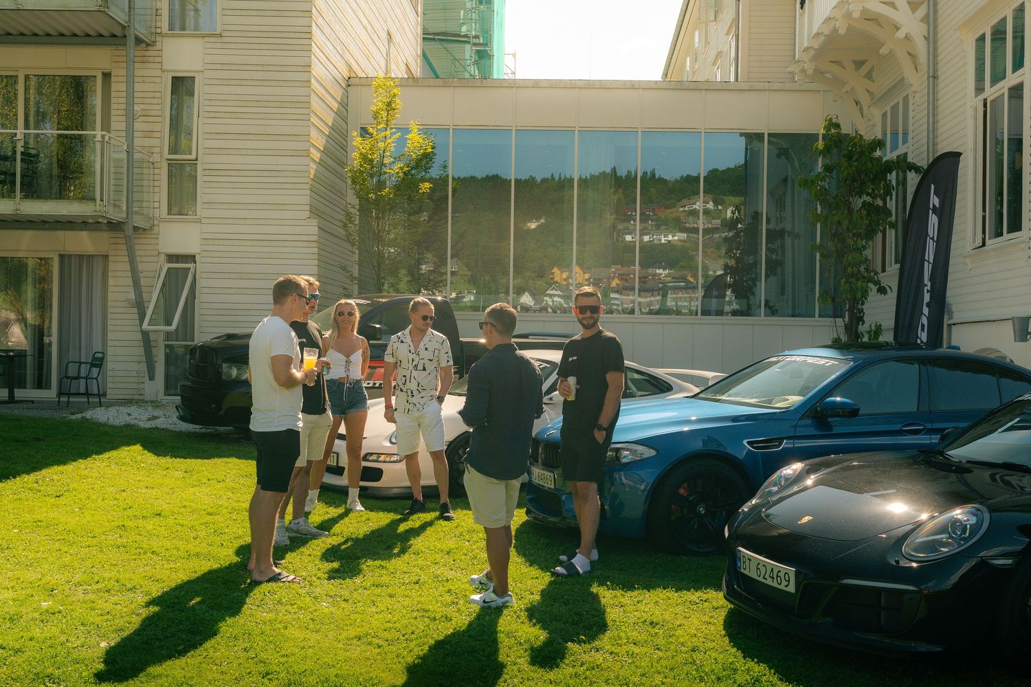 Group of people chatting on a sunny grass lawn beside parked sports cars outside a modern building with large glass windows.