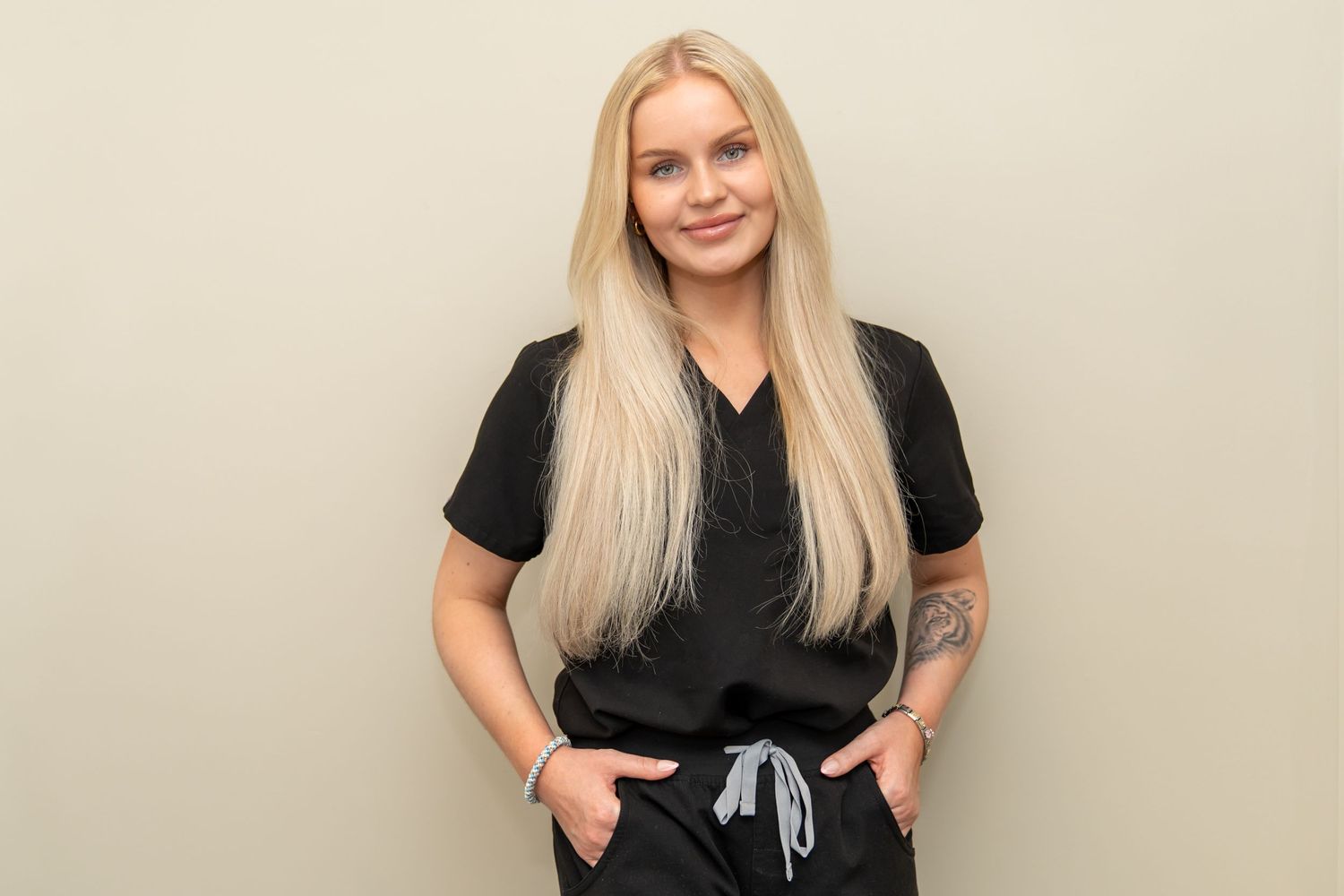 Smiling woman with long blonde hair wearing black scrubs, hands in pockets, standing against a neutral beige background.