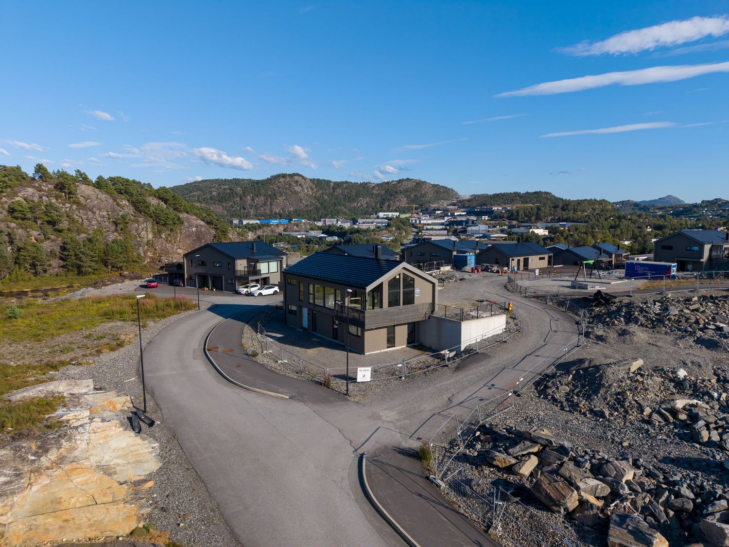 Aerial view of a modern wooden building beside a curved road in a rocky coastal development with hills and a clear blue sky.