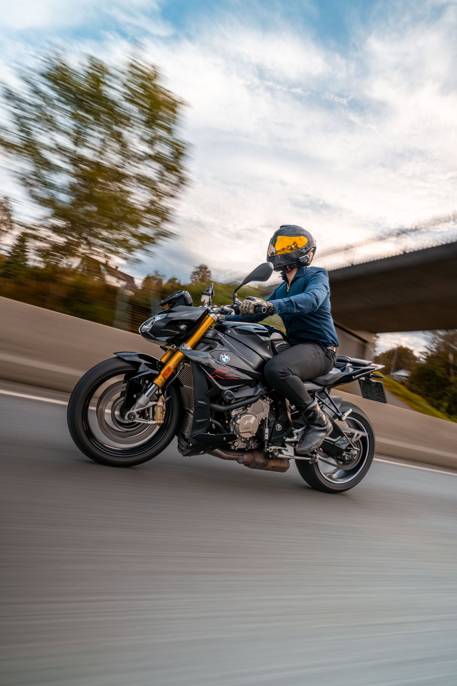 A motorcyclist rides a custom black and gold sports bike at speed down an open road against cloudy skies.
