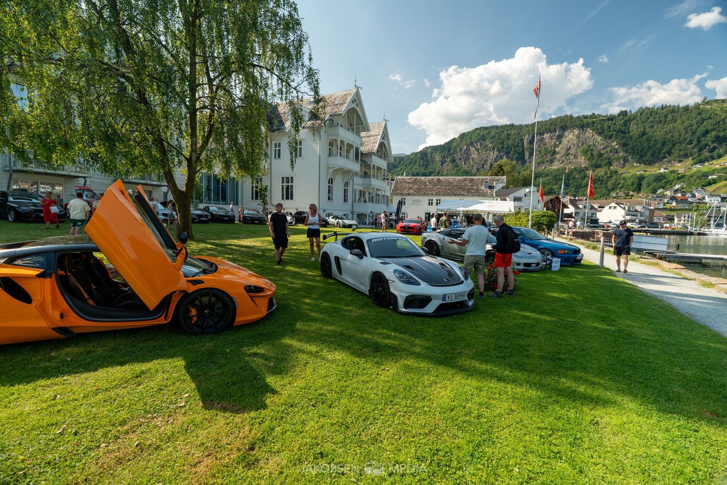 Supercar meetup on grassy lawn with orange McLaren and other luxury vehicles displayed.