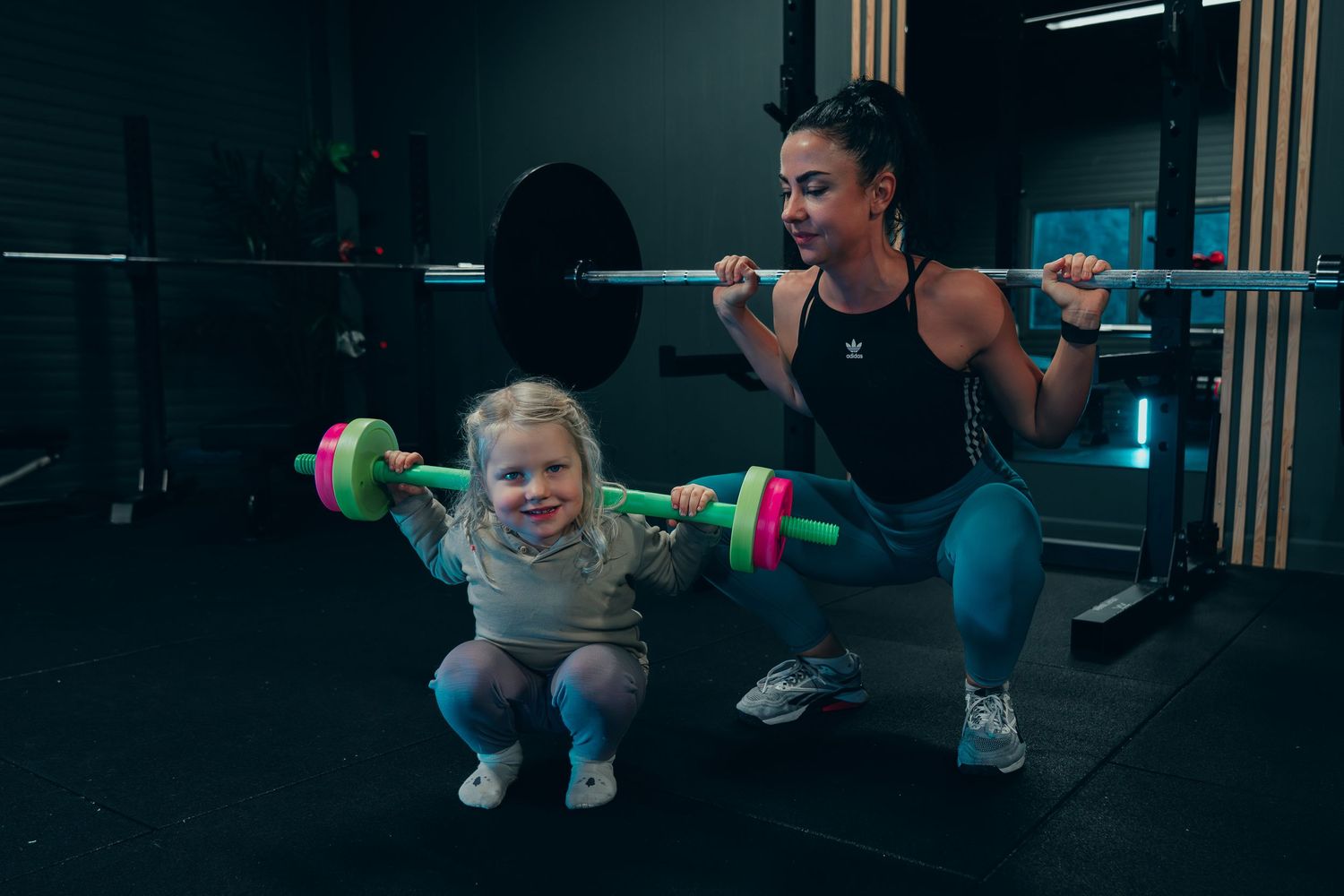 In a dimly lit gym, two people workout together, lifting colorful weights and barbells in synchronized form.